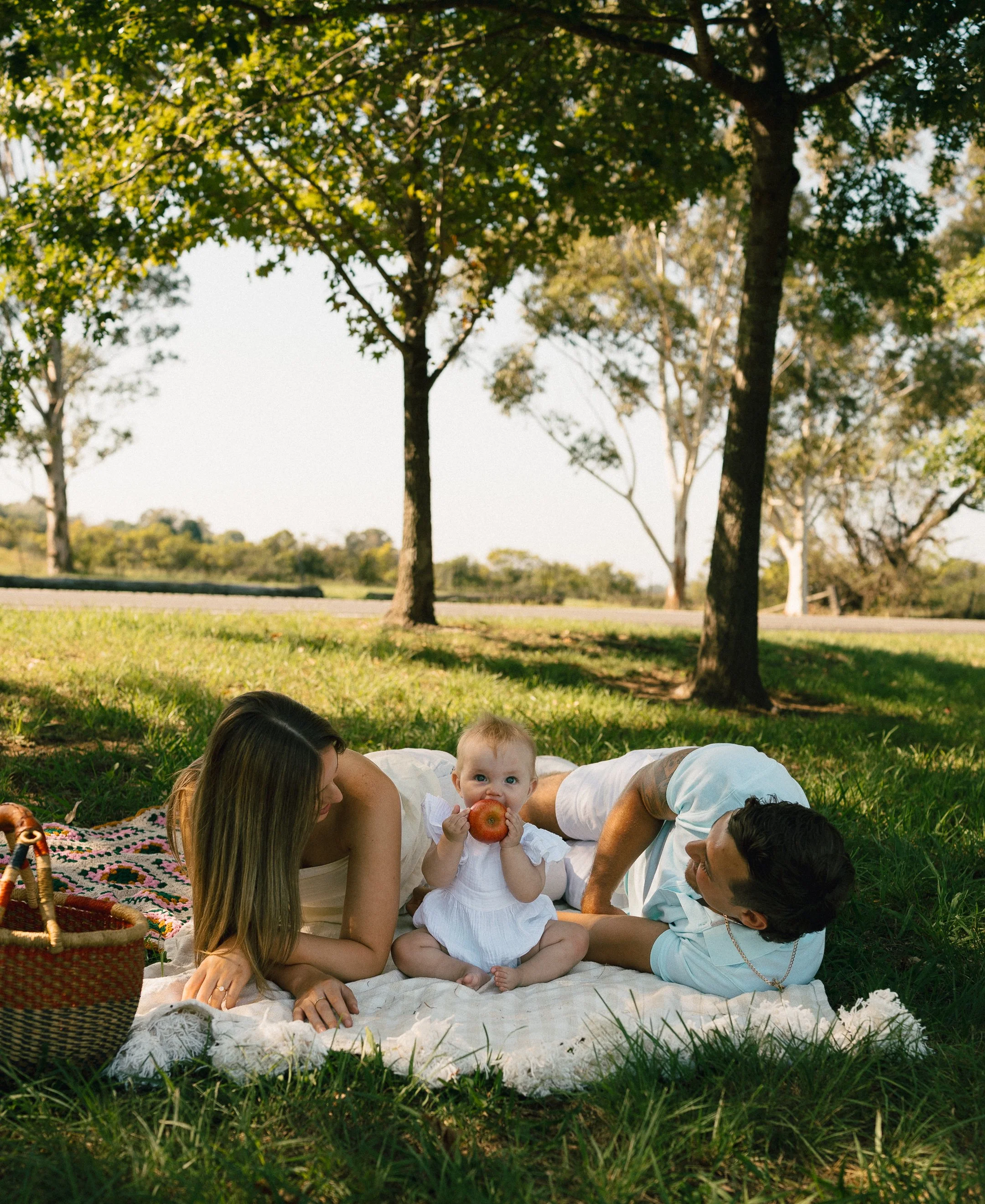 Family having a picnic outdoors, lying on a blanket under trees, with a child holding an apple and looking at the camera.