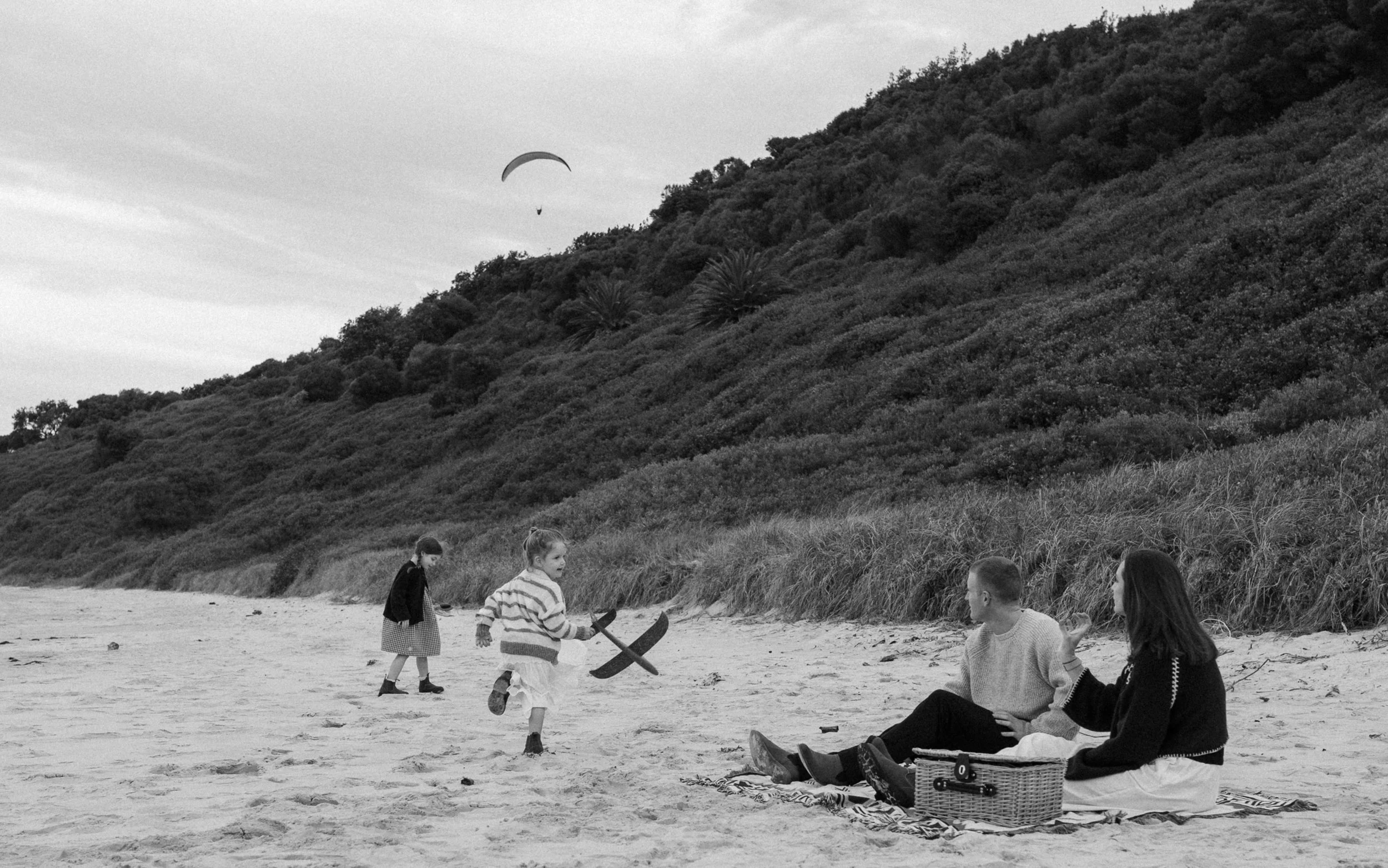 A black-and-white photo of children and a woman at the beach near a grassy hillside, with a person paragliding high in the sky.