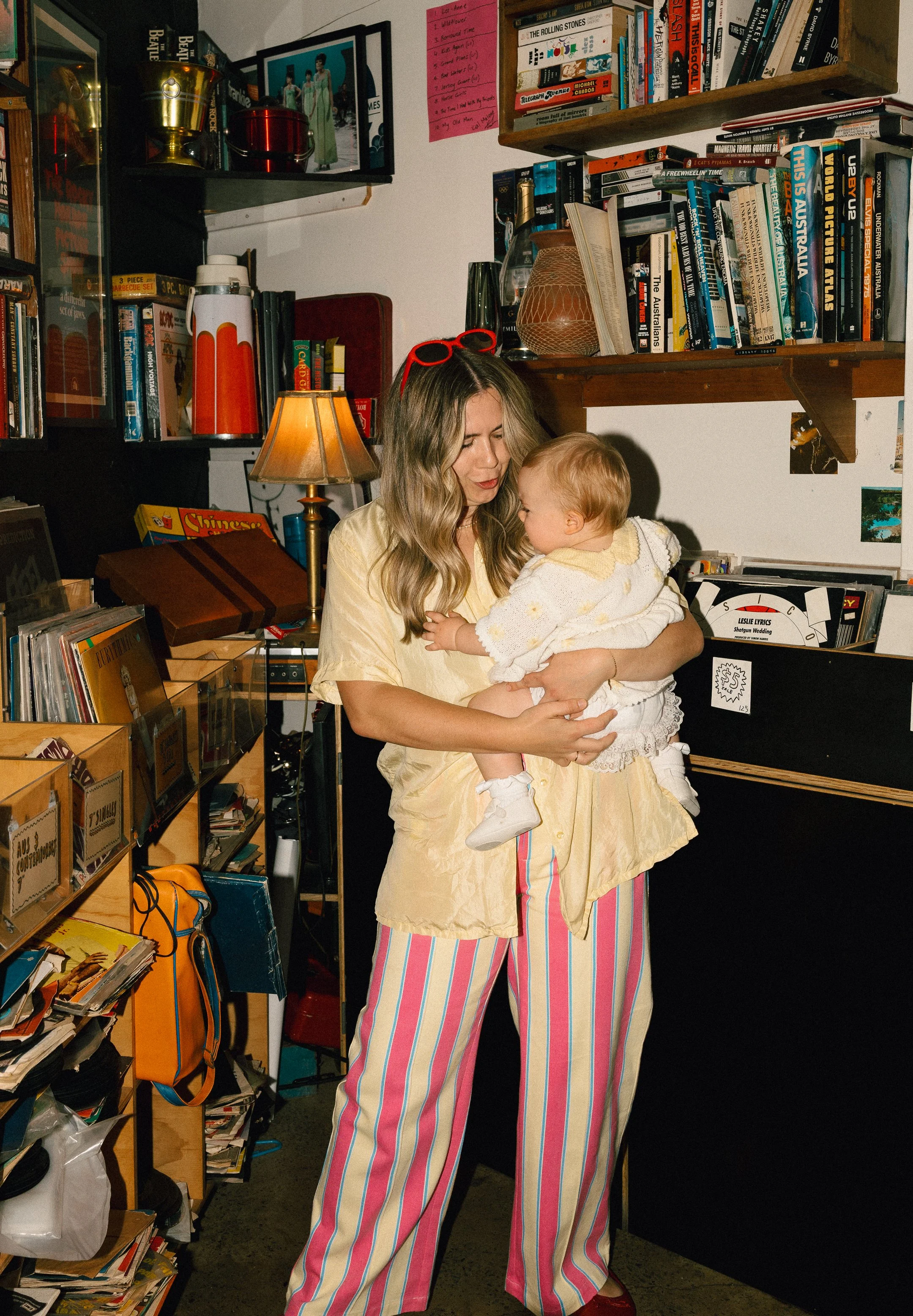 A woman holding a young girl in a room filled with books, magazines, and various objects, including a lamp, trophies, and colorful shelves. The woman is wearing yellow top and striped pants, and the girl is in a light-colored dress with a yellow collar.