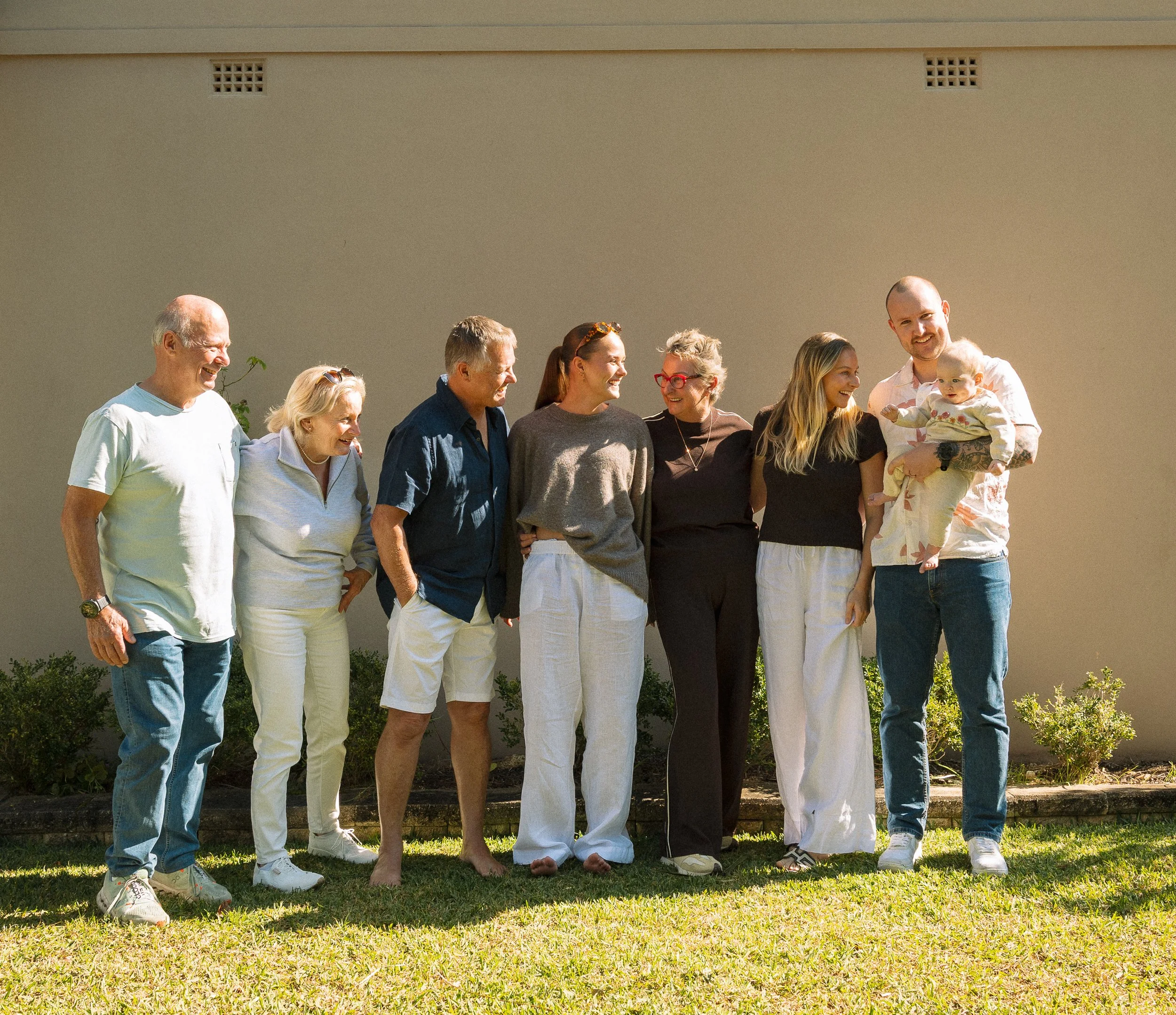 A group of people standing outdoors in a line, smiling, with a plain beige wall in the background.