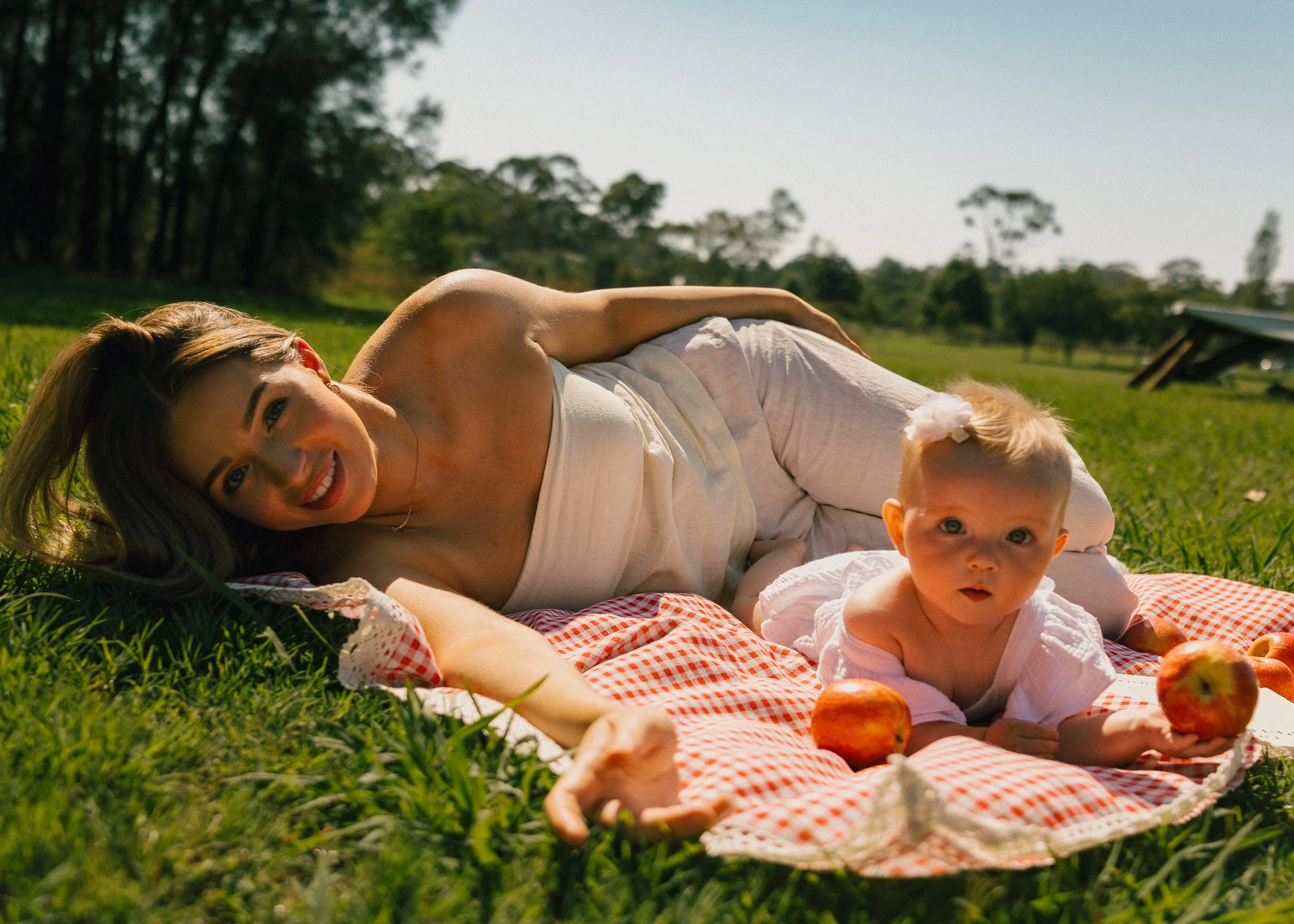 A woman and a baby lying on a red and white checkered blanket outdoors on grass, with apples around them and trees in the background.