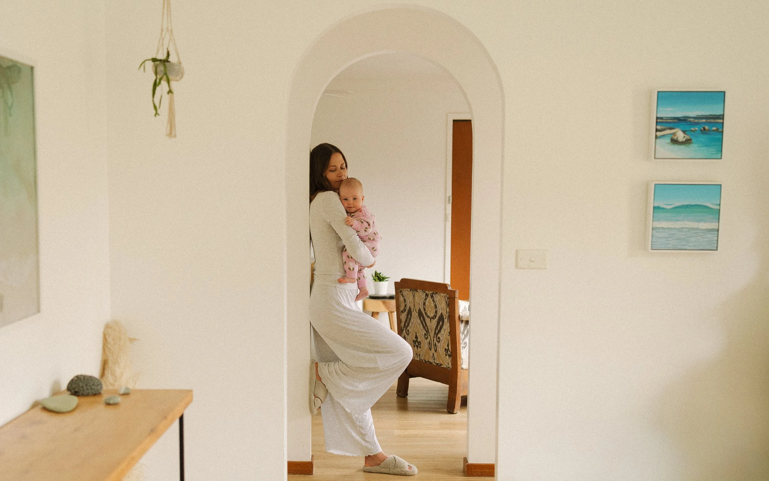 A woman in white pajamas holding a baby in pink pajamas with hearts inside a home, seen through an arched doorway.