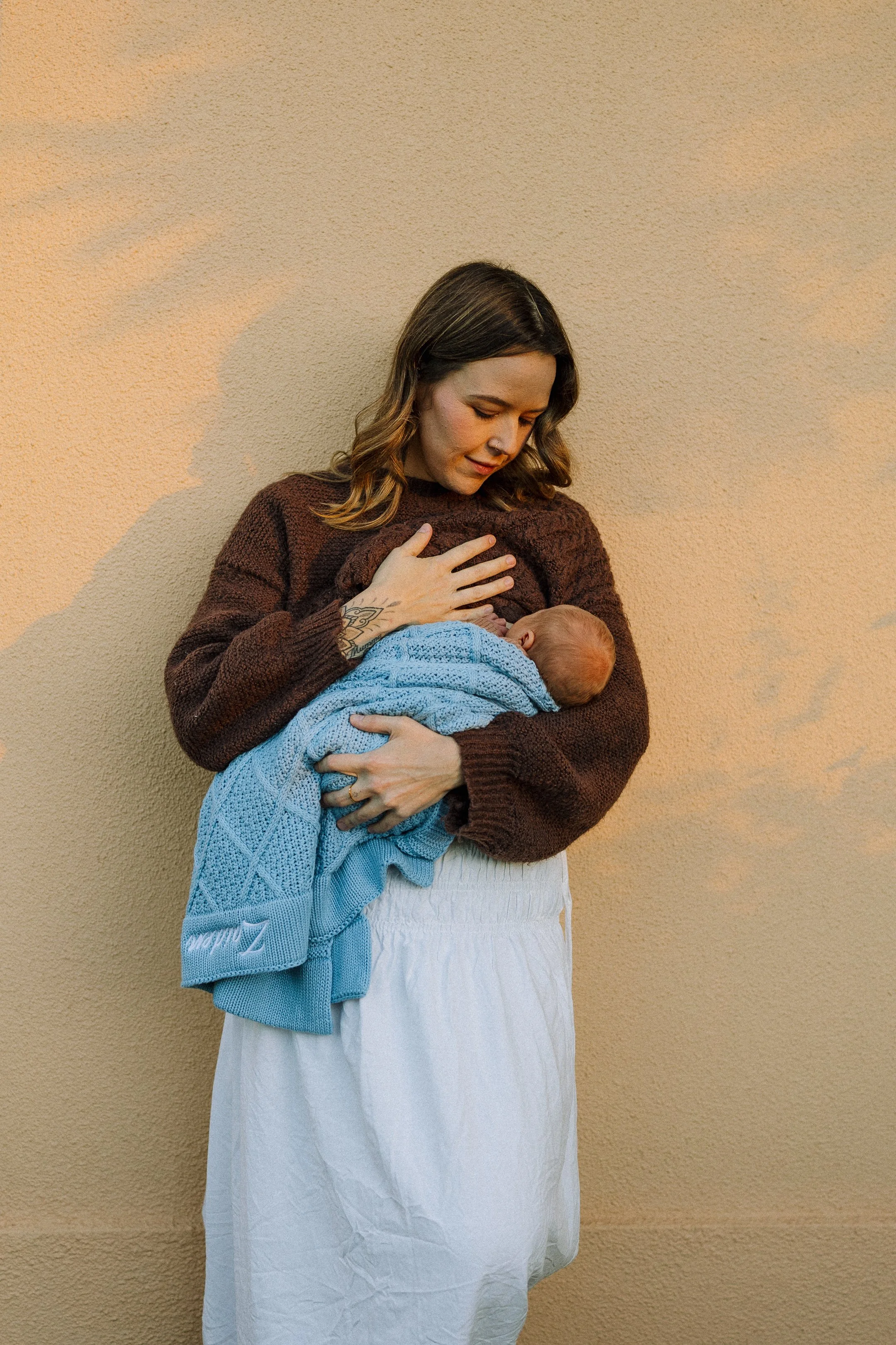 A woman in a brown sweater holding a newborn wrapped in a blue blanket, standing against a beige wall.