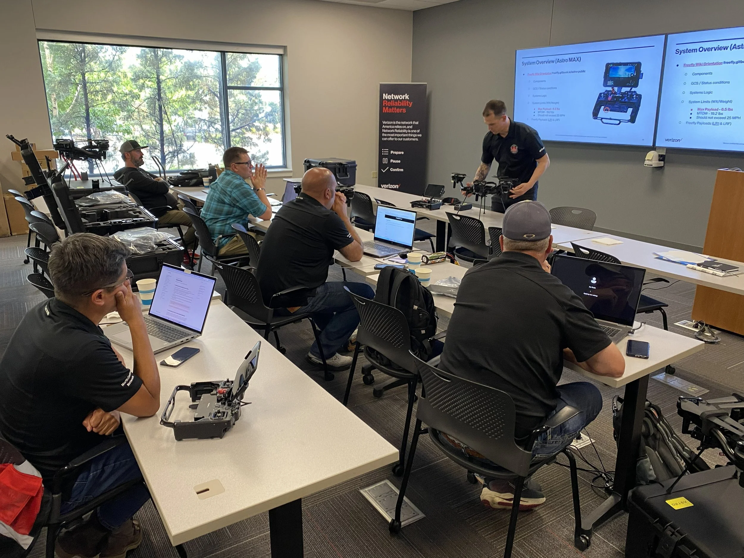 A group of students attending a classroom presentation in a conference room.  An instructor is set up with drones and cameras at the front, and a large screen displaying presentation slides.