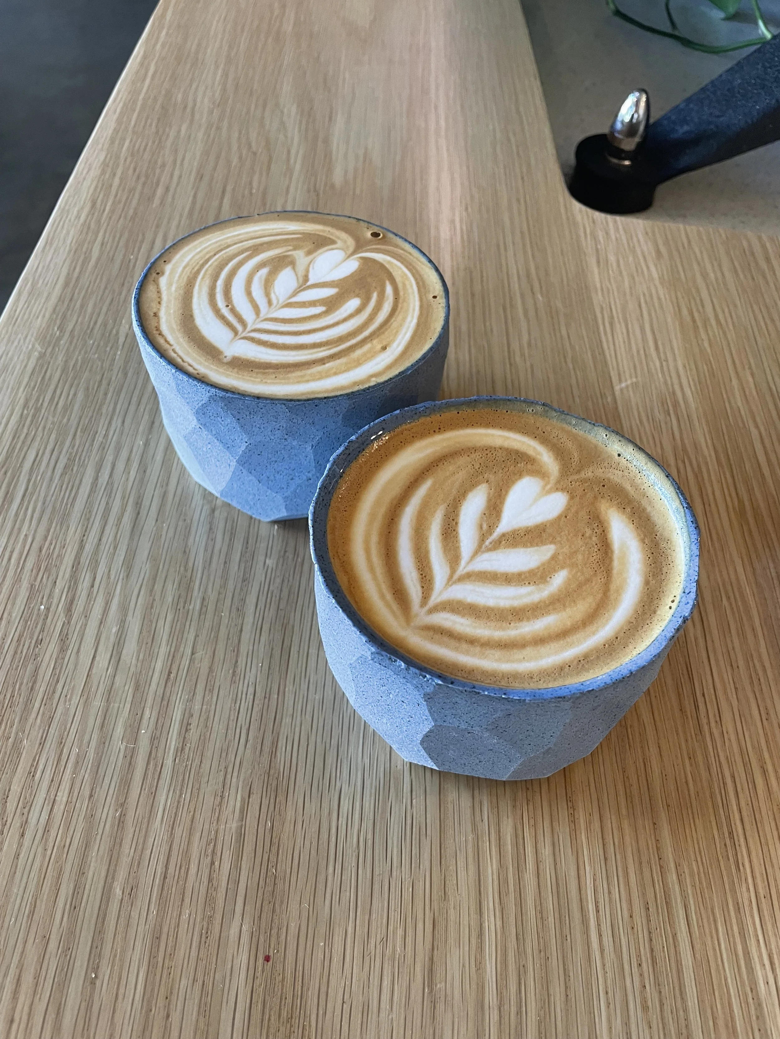 Two cups of coffee with latte art on a wooden table, one in a gray cup and the other in a blue cup.