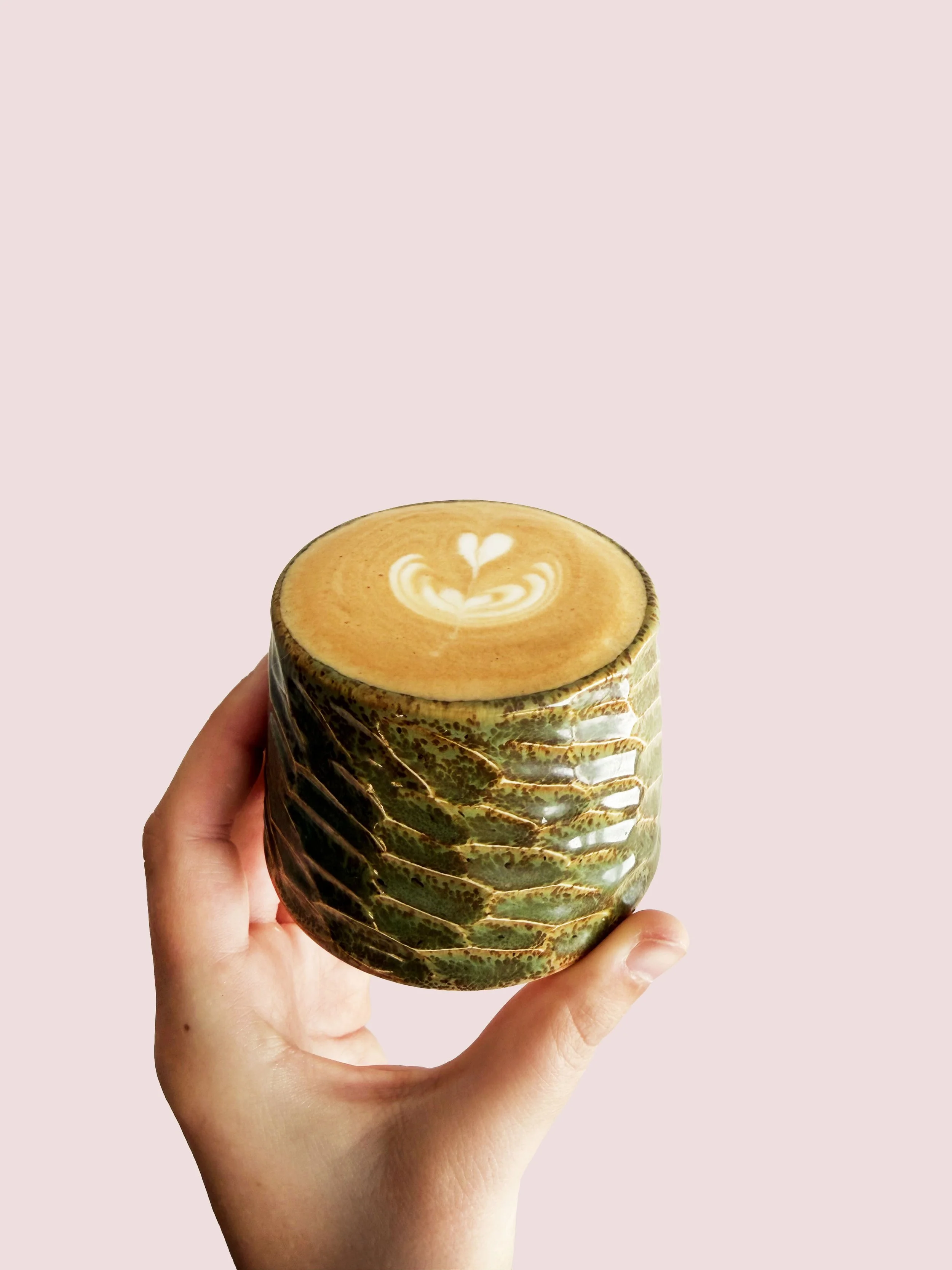 A hand holding a ceramic cup with latte art in the shape of a heart on top, against a pastel pink background.
