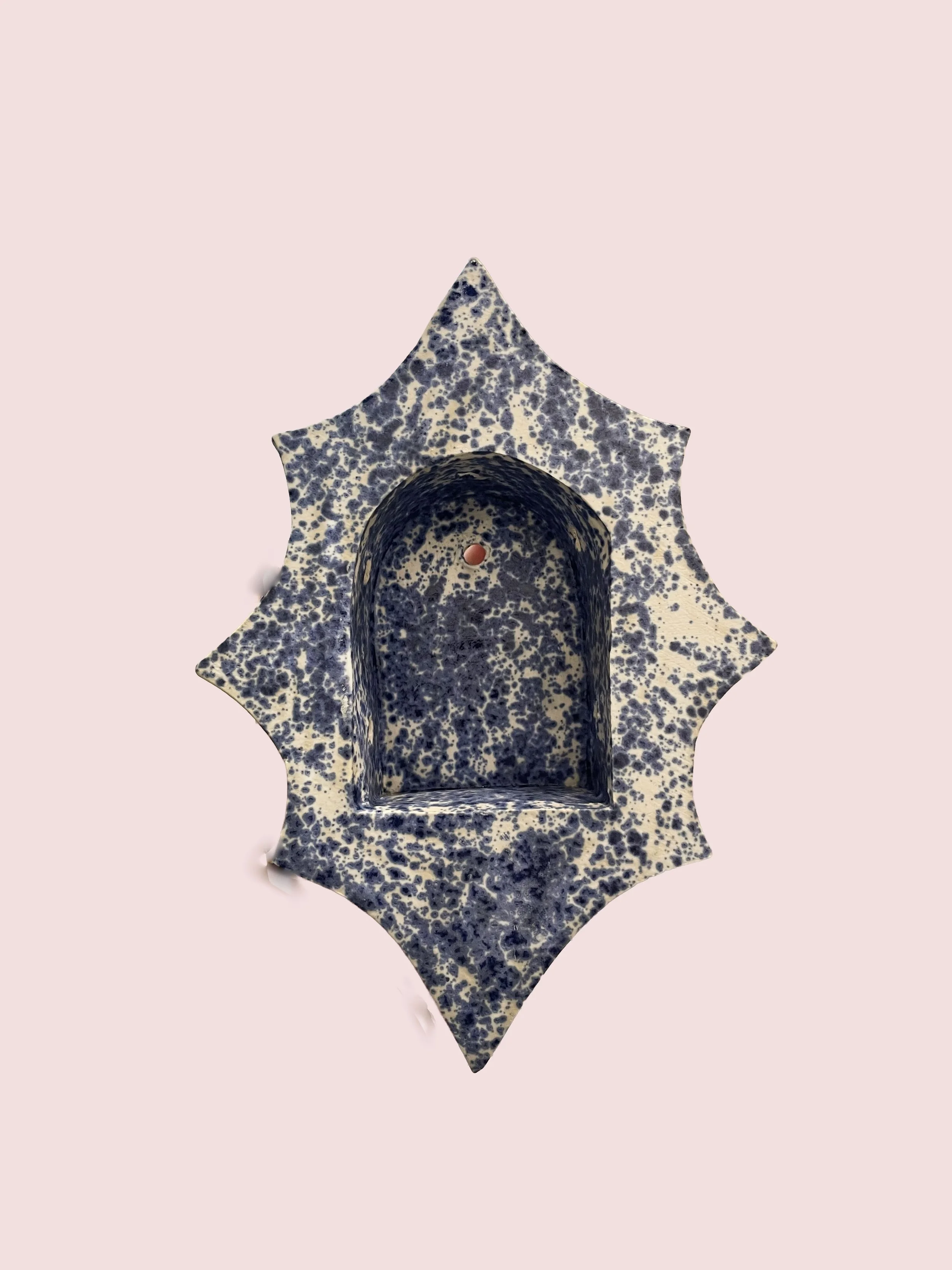 A ceramic star-shaped stand with a speckled dark blue and cream pattern, featuring a round hollow in the center, placed against a pale pink background.