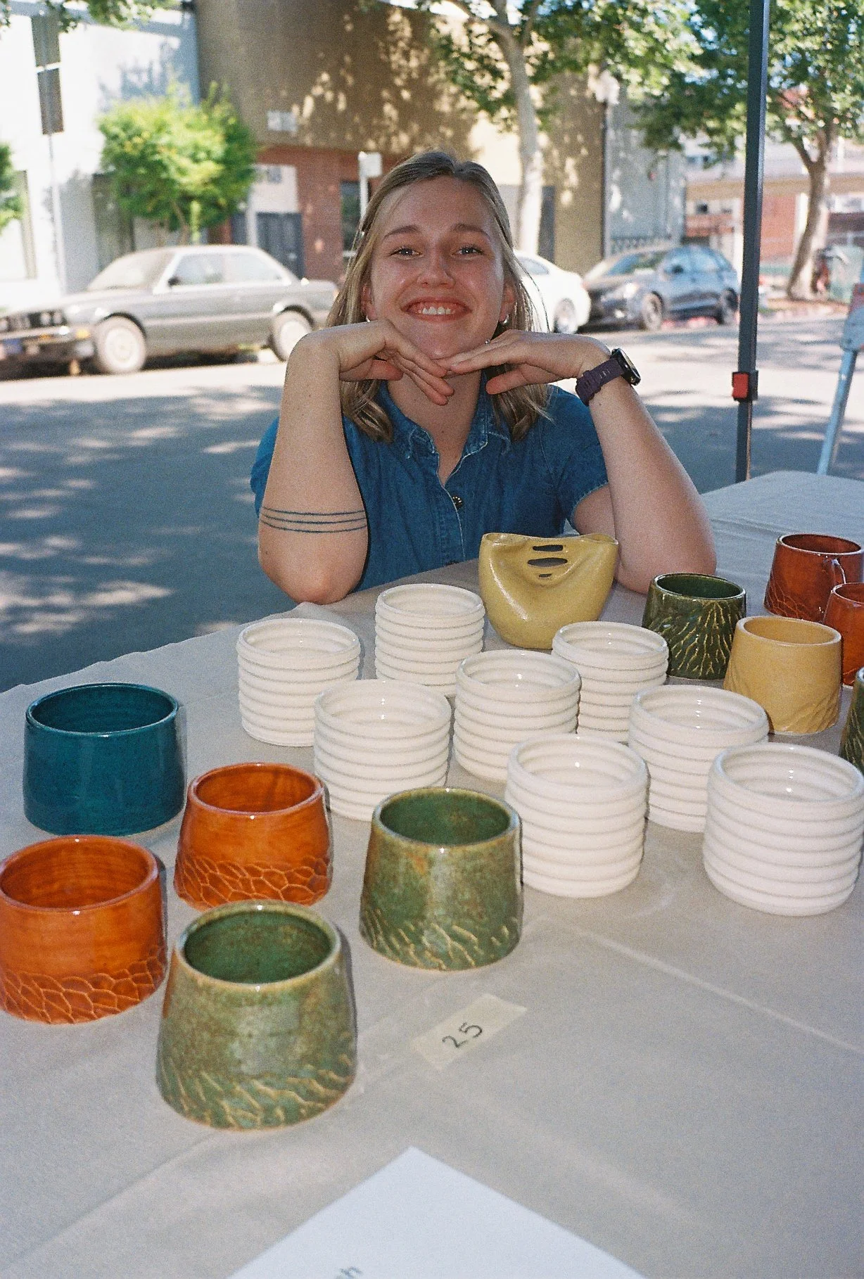 A woman sitting at a table with various colorful pottery cups and bowls on display, outdoors on a sunny day.