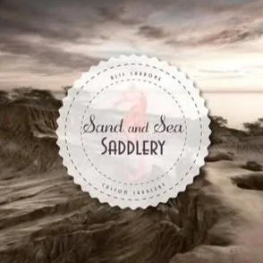 A logo with a beach scene and cliffs in the background, reading "Sand and Sea Saddlery".