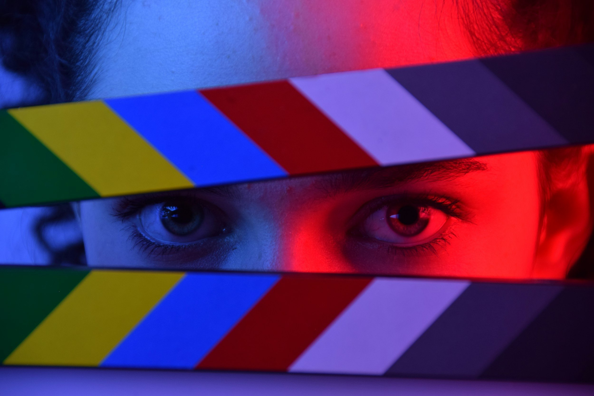 Close-up of a child's face with only the eyes visible, looking through a striped, colorful film clapper with blue, yellow, red, purple, and white stripes, illuminated by red and blue lighting.