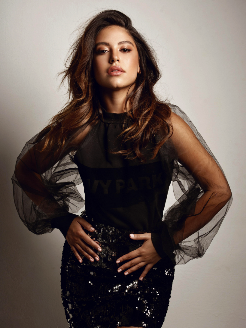 A woman with long, wavy brown hair wearing a black top with sheer, puffed sleeves and a sequined black skirt posing confidently against a neutral background.