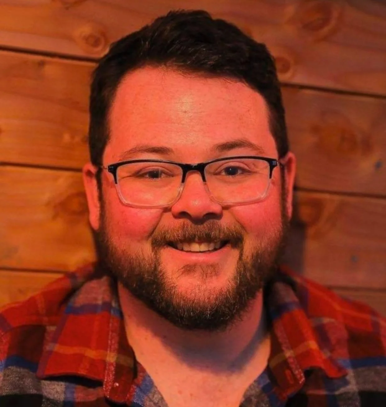 A smiling man with glasses and a beard wearing a red plaid shirt, standing in front of a wooden wall.