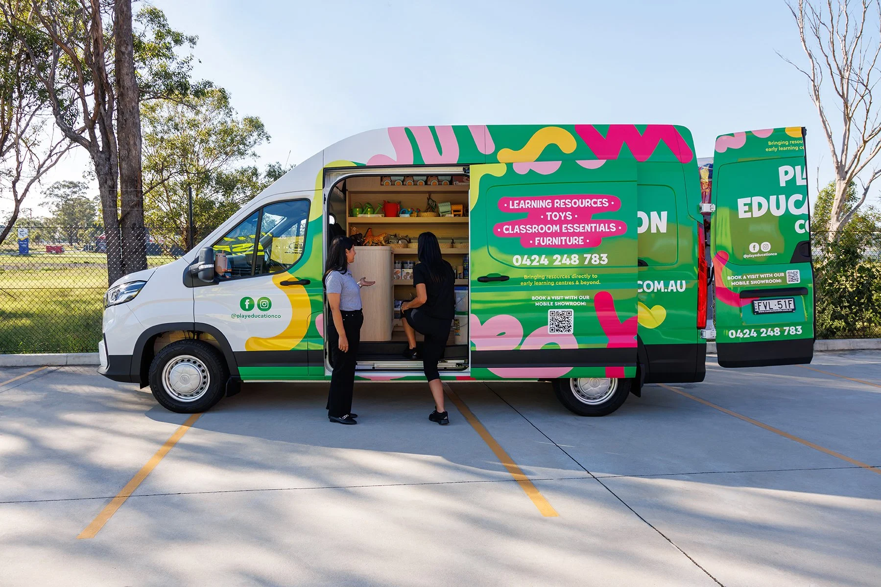A mobile education van parked in a lot with two women talking near an open side door, displaying various toys and classroom essentials on shelves inside.