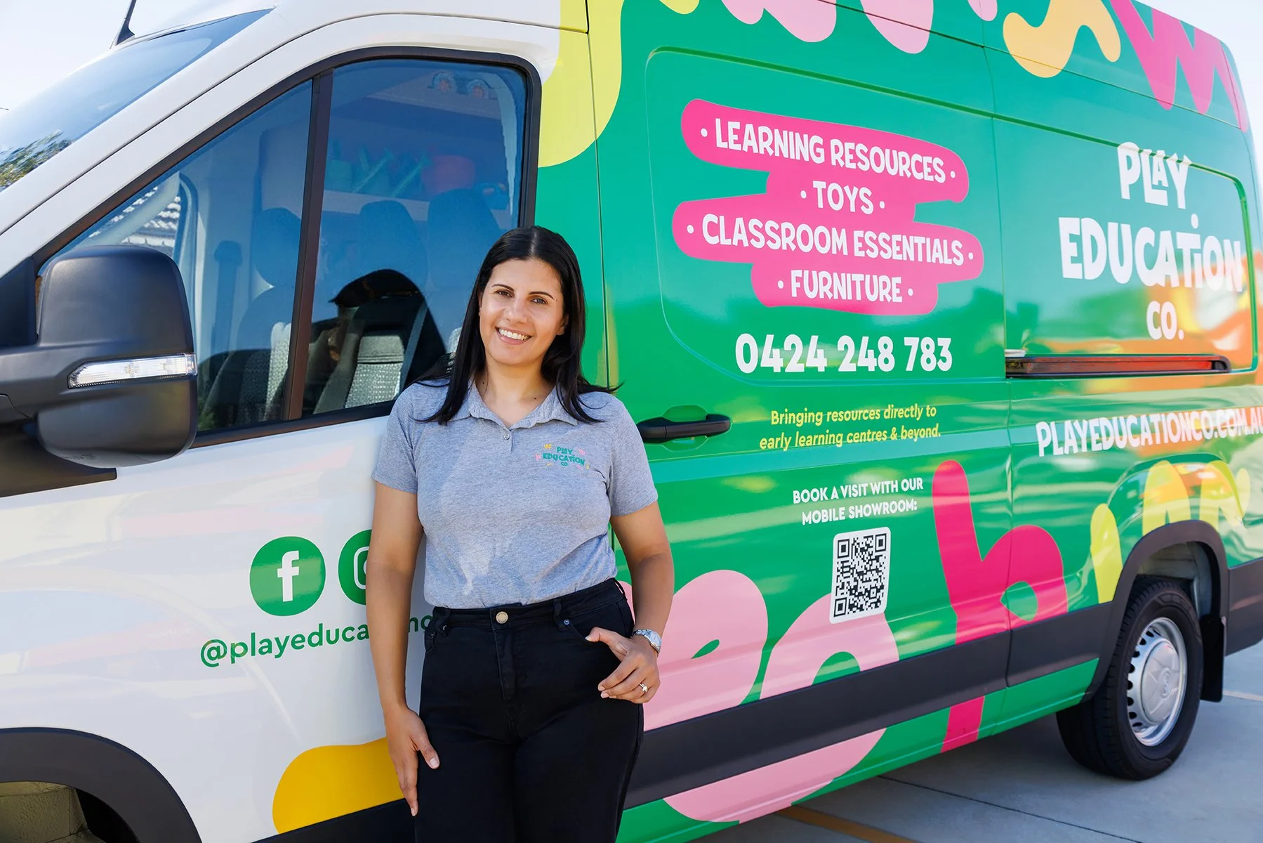 A smiling woman in a gray polo shirt standing in front of a colorful van with vibrant green, pink, and yellow graphics. The van has text promoting educational resources, toys, classroom essentials, and furniture. There are social media icons, a QR code, and a phone number on the van.