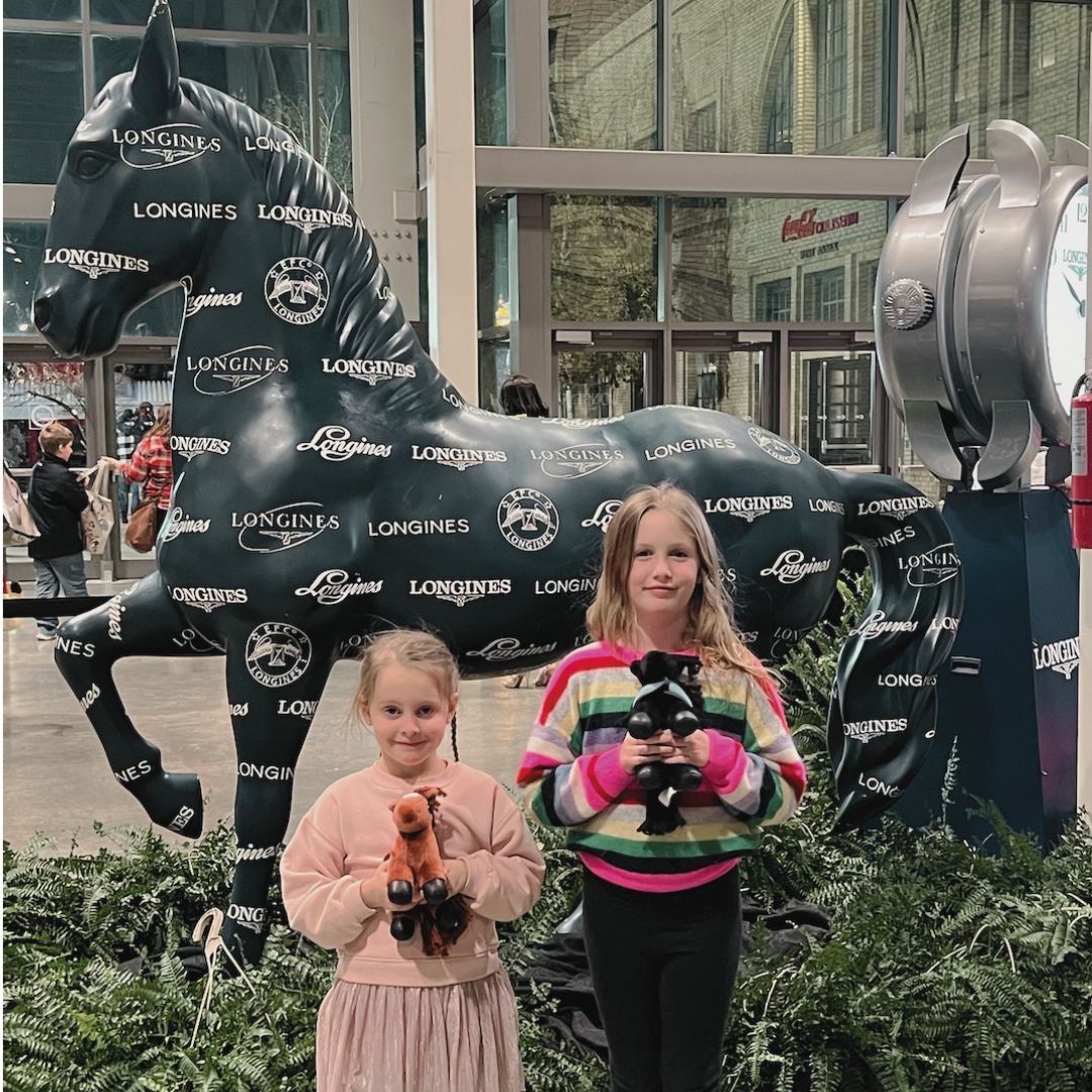 Two young girls standing in front of a large black horse-shaped display covered with Longines brand logos, holding plush animal toys.