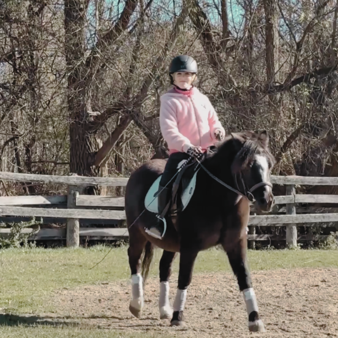 A young girl wearing a pink hoodie and a black helmet riding a black and white pony in an outdoor riding area with a wooden fence and leafless trees in the background.
