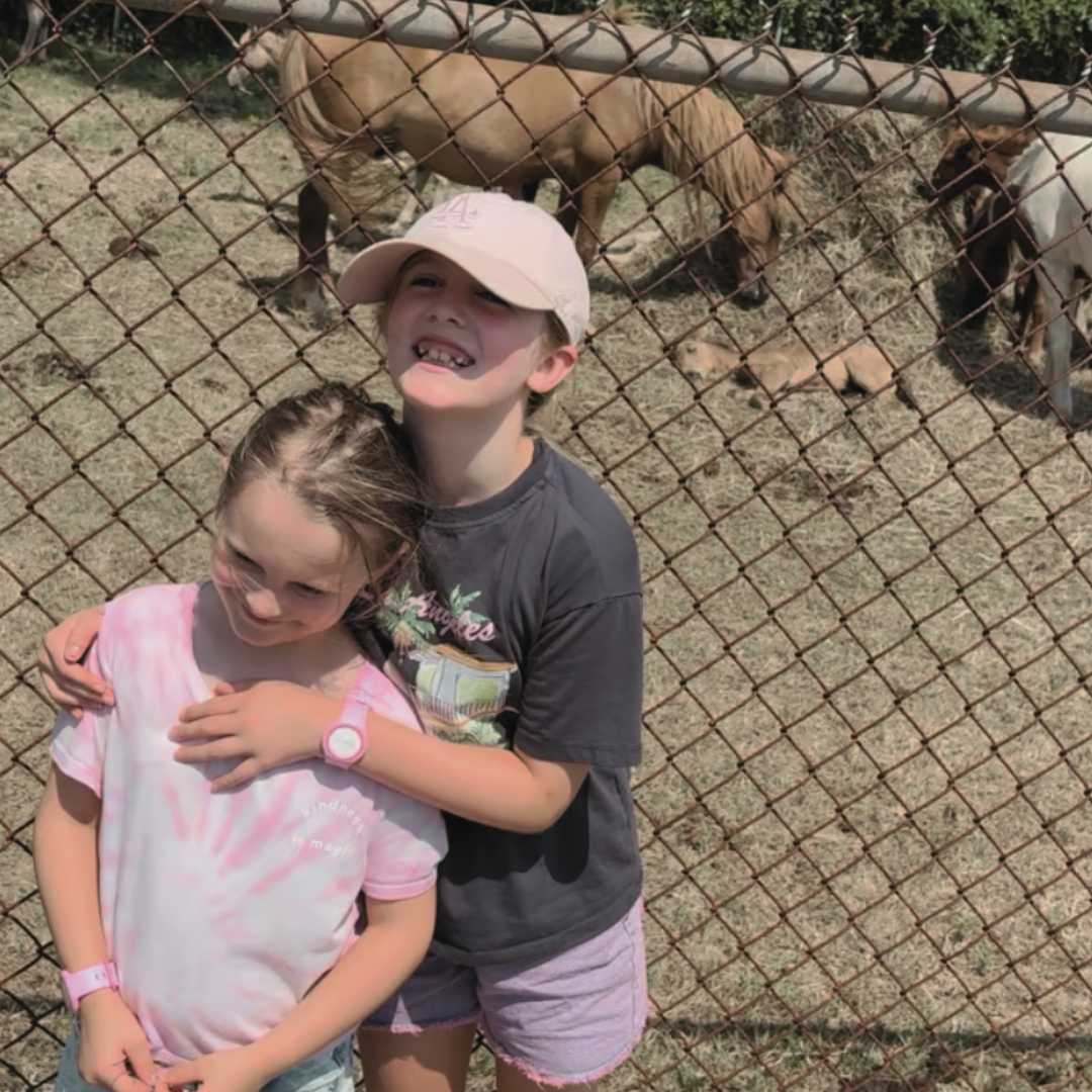 Two young girls hugging in front of a horse enclosure, with a brown and a white horse visible behind the fence.