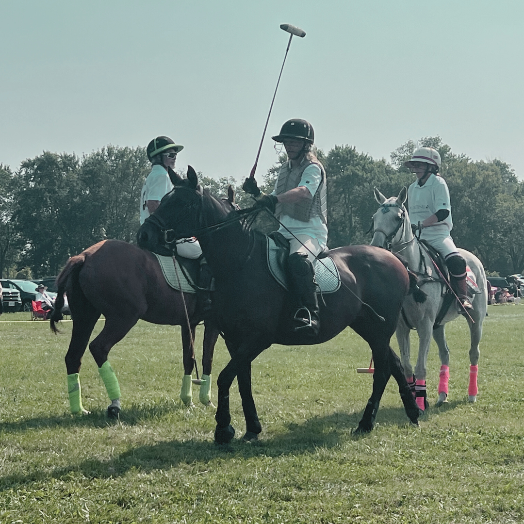 Three polo players on horseback on a grassy field, one holding a polo mallet, with trees and parked cars in the background.