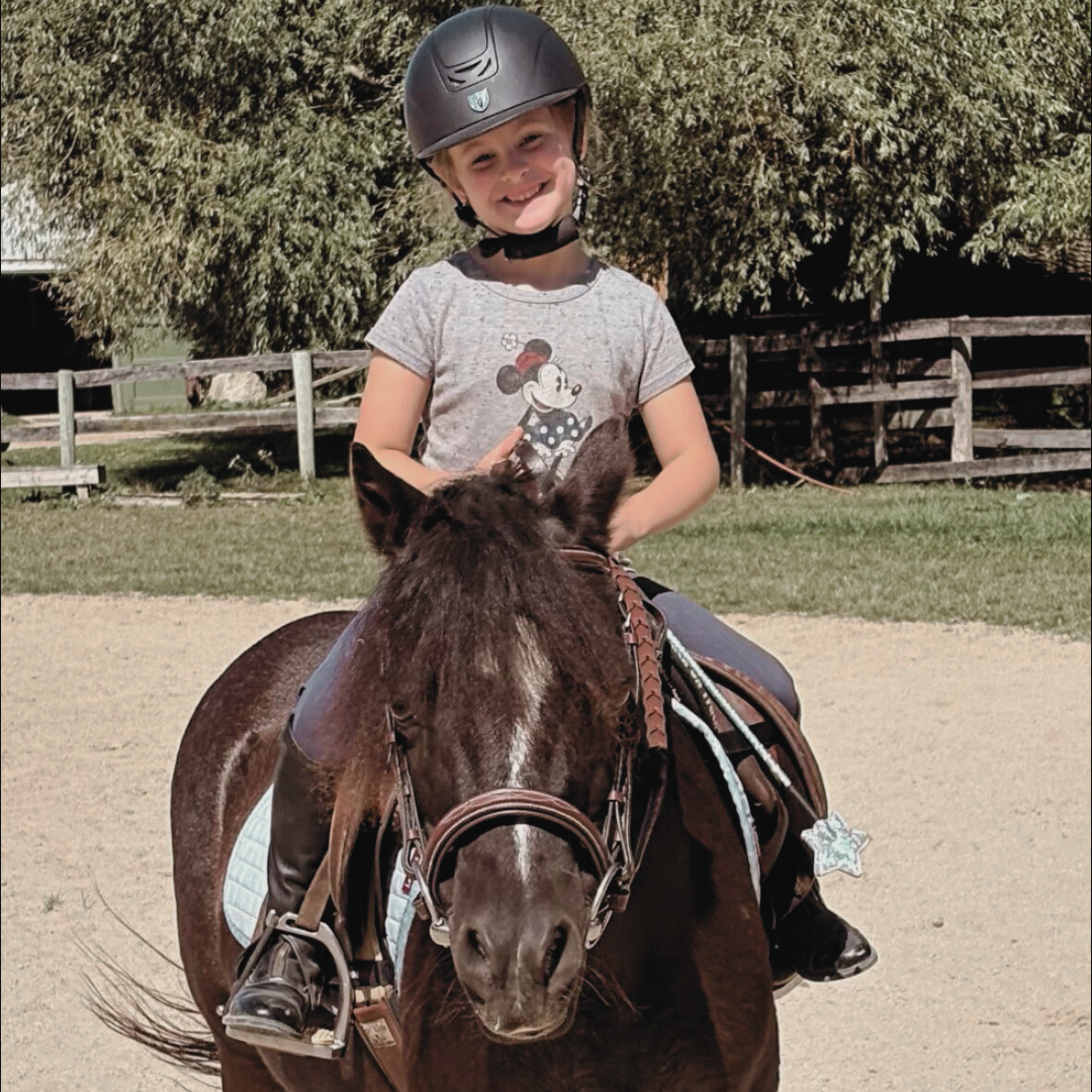 A young girl riding a brown horse outdoors, wearing a black riding helmet and a Minnie Mouse t-shirt, smiling.