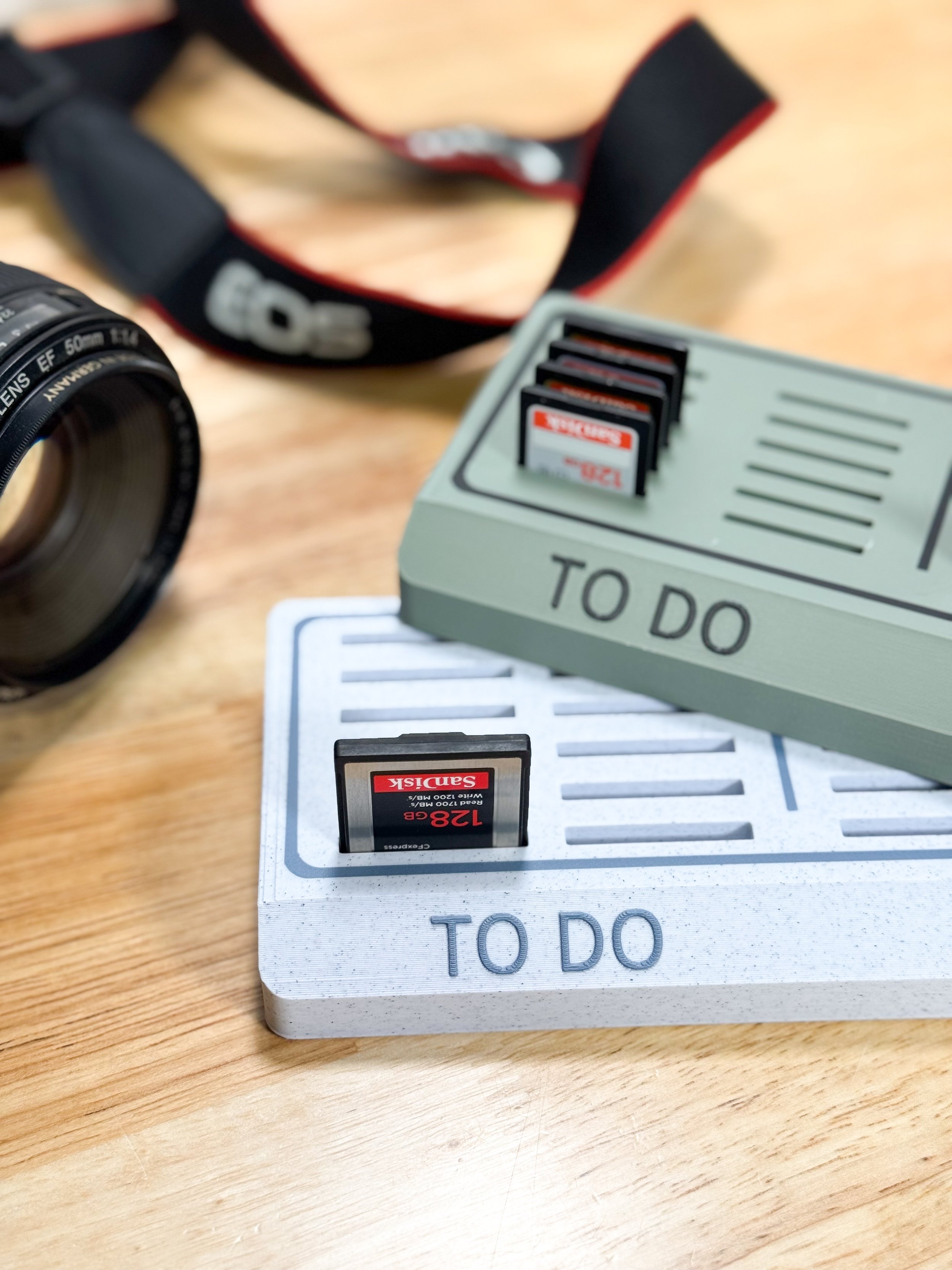A camera lens, a camera strap, and two storage cases labeled 'TO DO' with SD cards, one inside each case, on a wooden surface.
