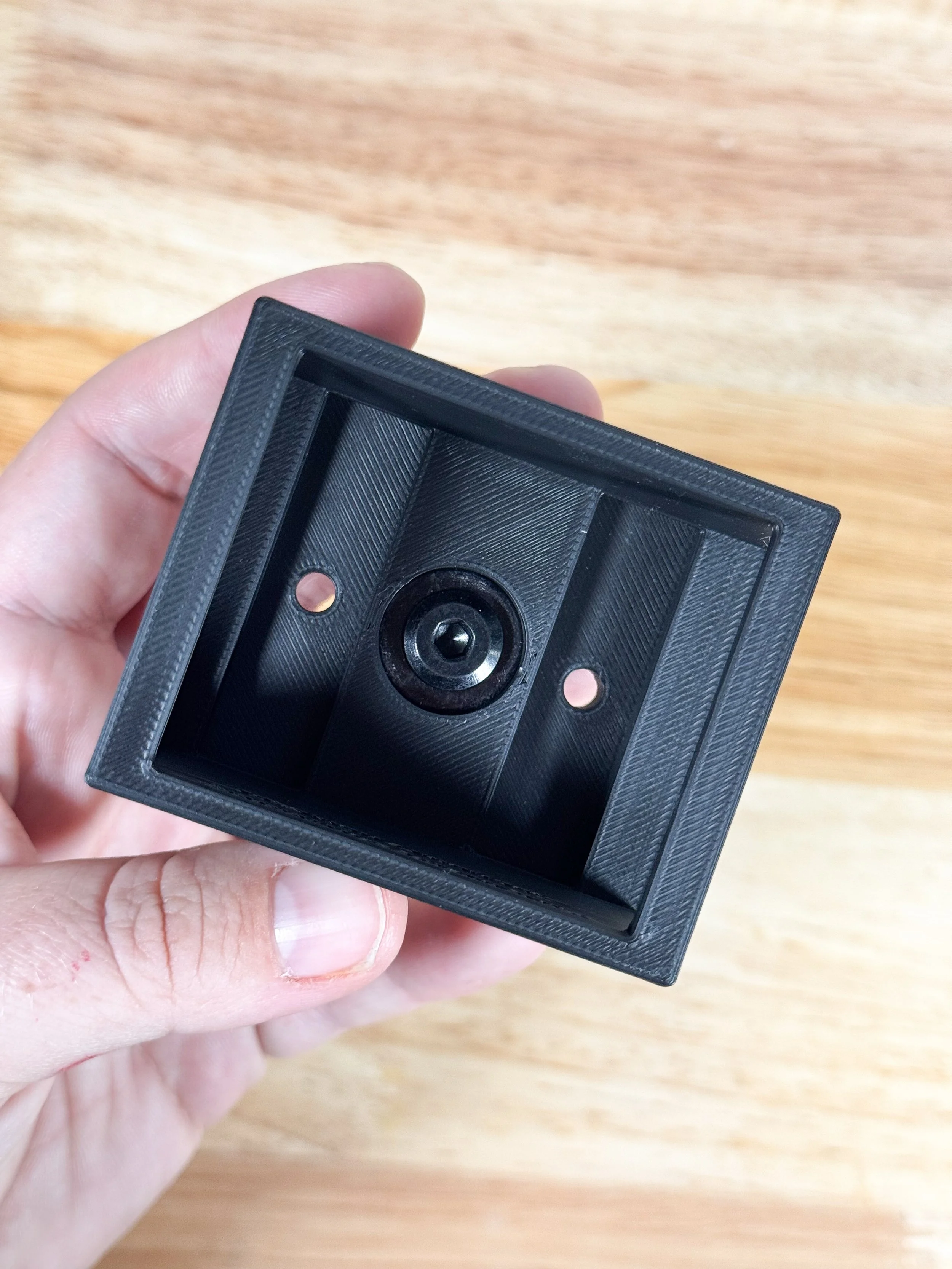 A black, square-shaped wall mount with a circular opening in the center, held in a person's hand against a wooden surface background.