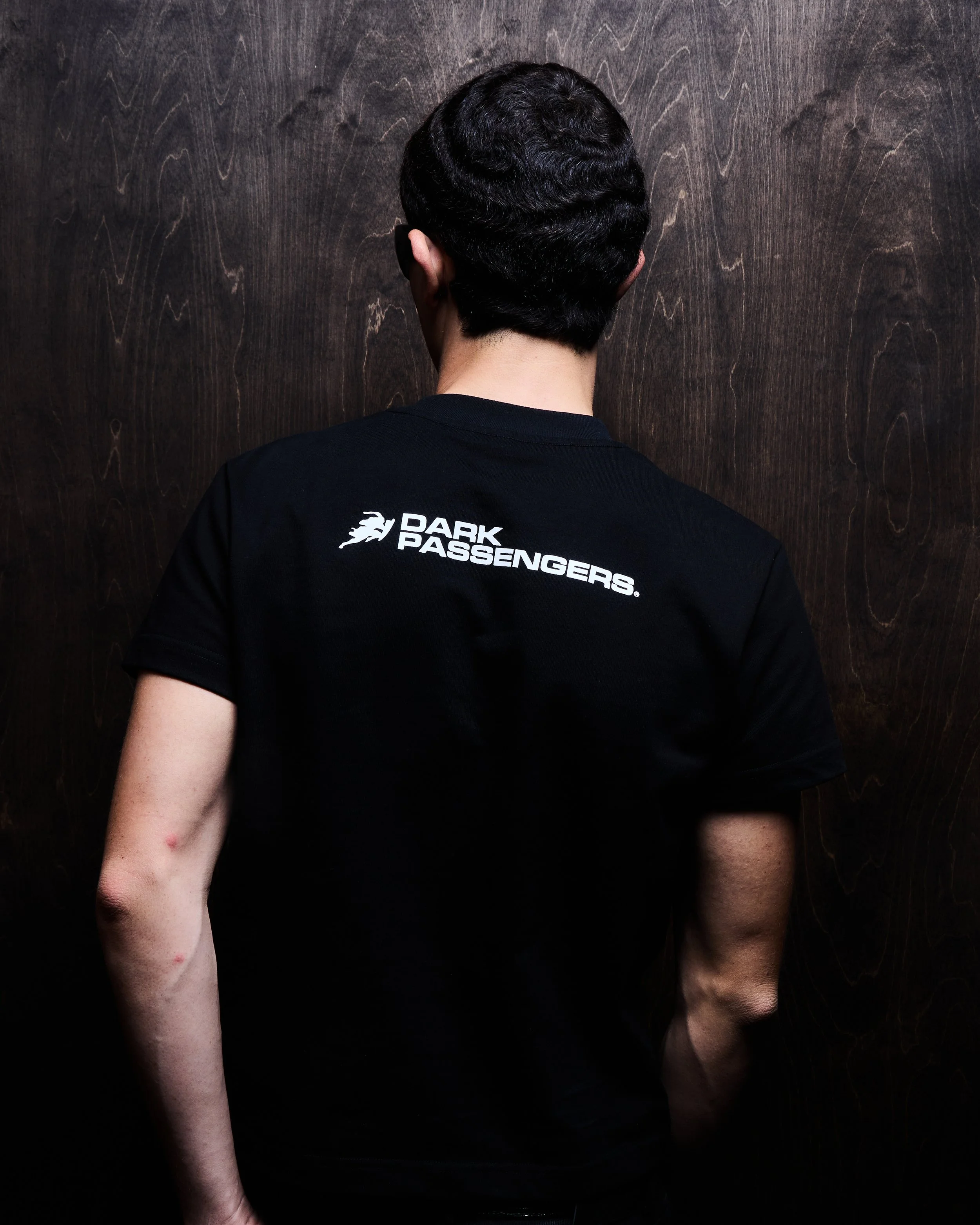Person with dark hair wearing a black T-shirt with 'Dark Passengers' logo on the back, standing against a dark wooden wall.