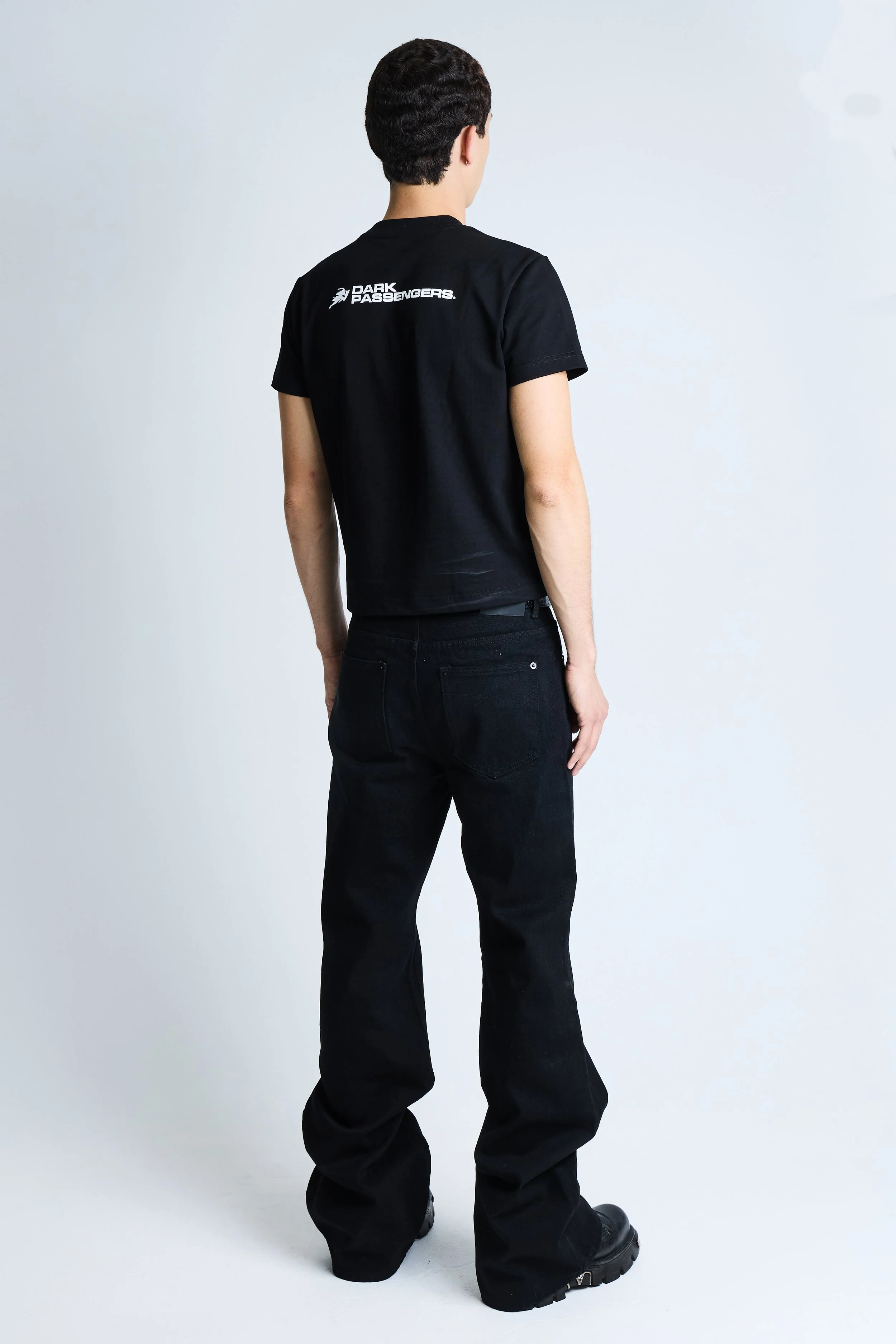 Back view of a person wearing a black t-shirt with white text and logo, black jeans, and black shoes, standing against a plain white background.