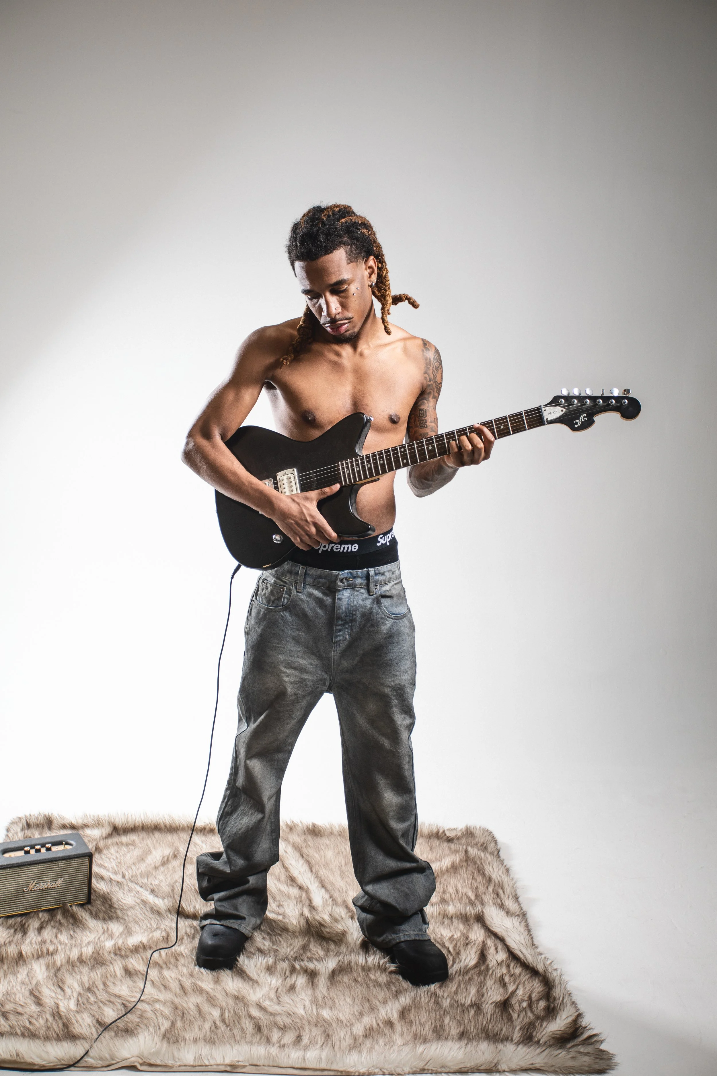 A shirtless man with dreadlocks playing an electric guitar on a faux fur rug, with a small amplifier nearby.