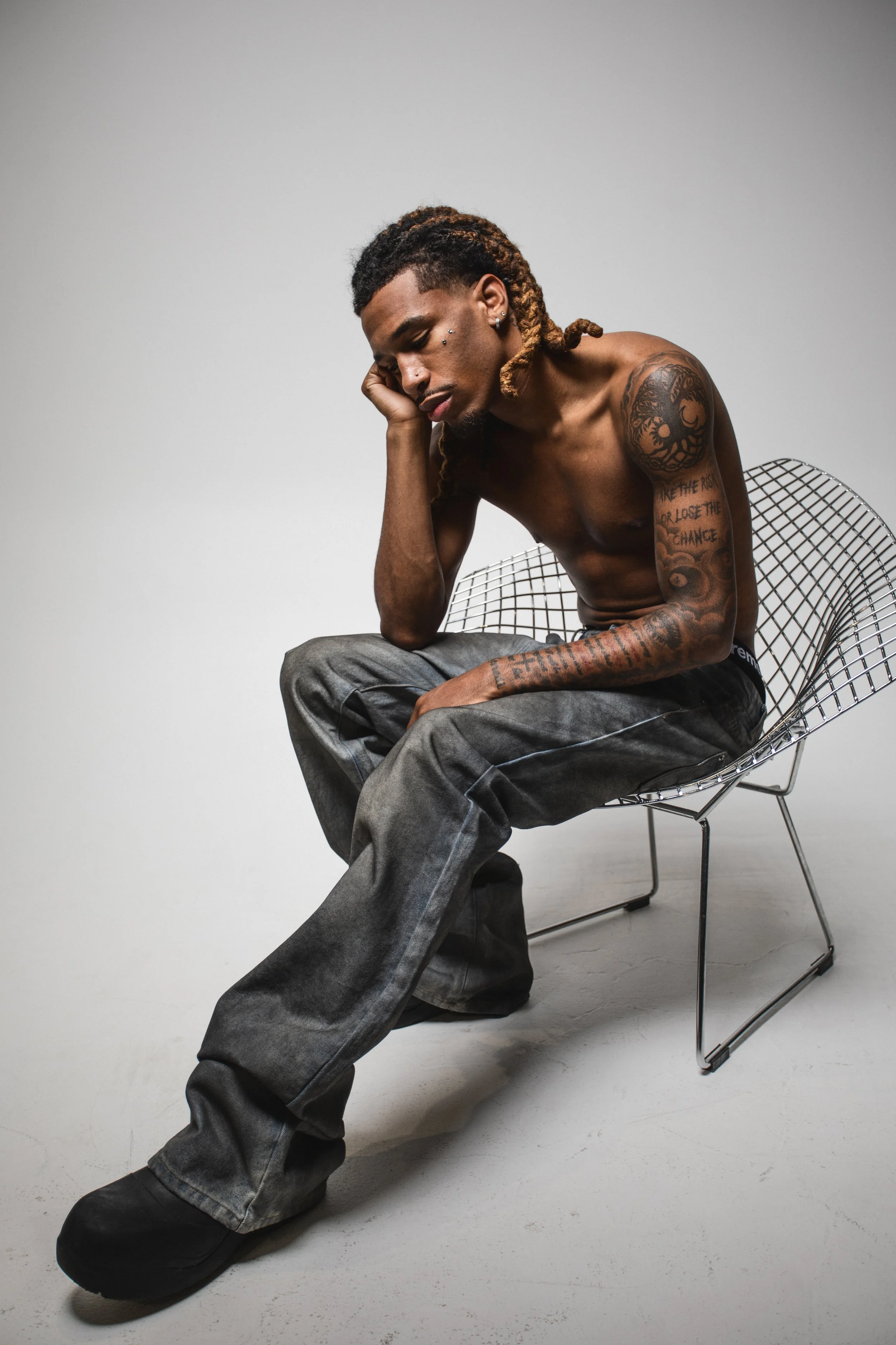 A shirtless man with tattoos sitting on a modern wire chair, resting his head on his hand, wearing loose gray jeans and black shoes, in a minimalist studio setting.