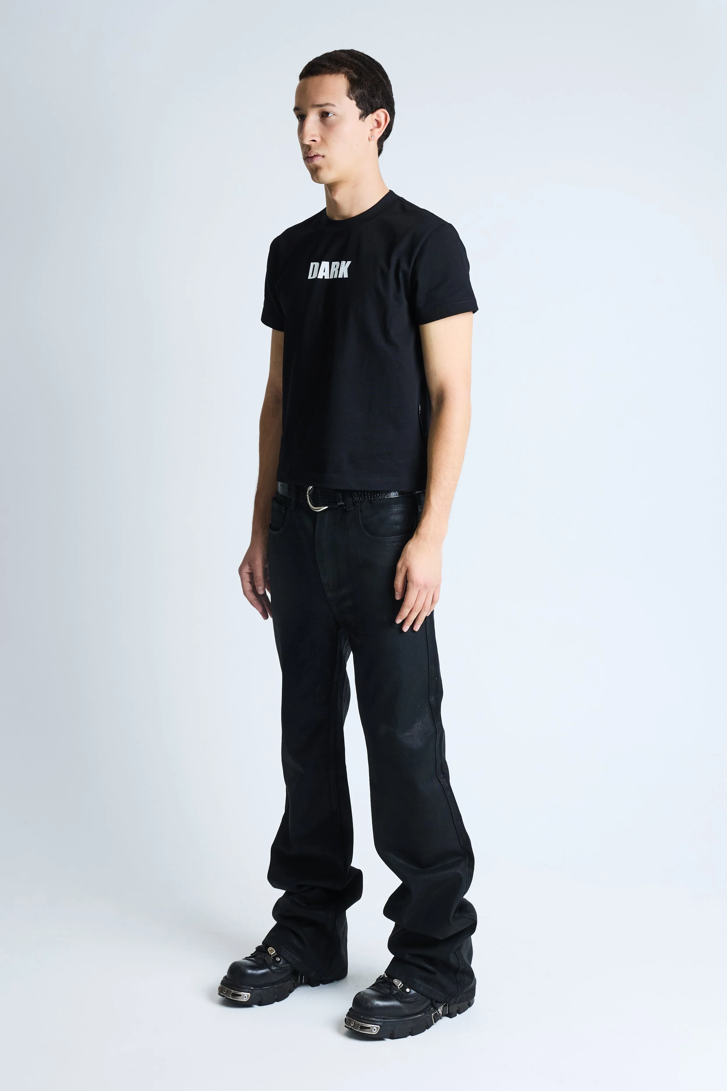 Young man with short dark hair, dressed in a black T-shirt with 'DARK' printed on it and black pants, standing against a plain white background.