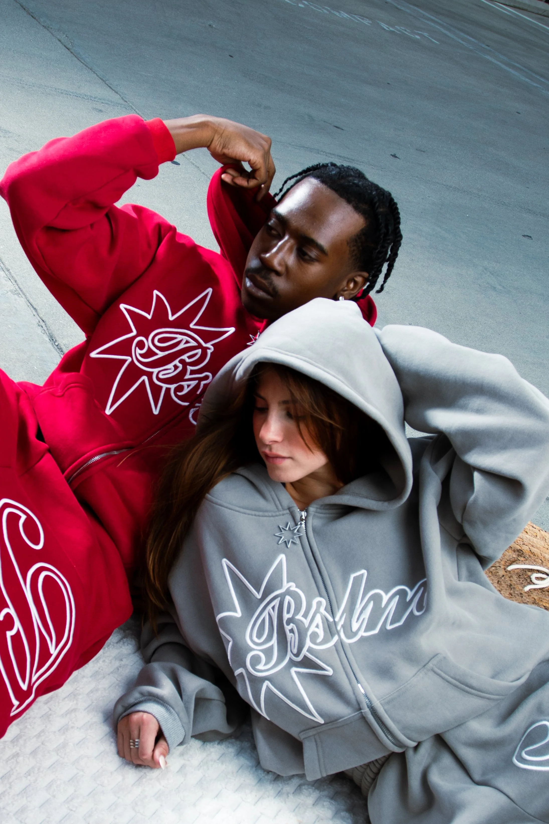 Two young adults, a man and a woman, are lying on the ground next to a curb, wearing hoodies with graphic designs. The man is in a red hoodie, and the woman is in a gray hoodie with the hood up.