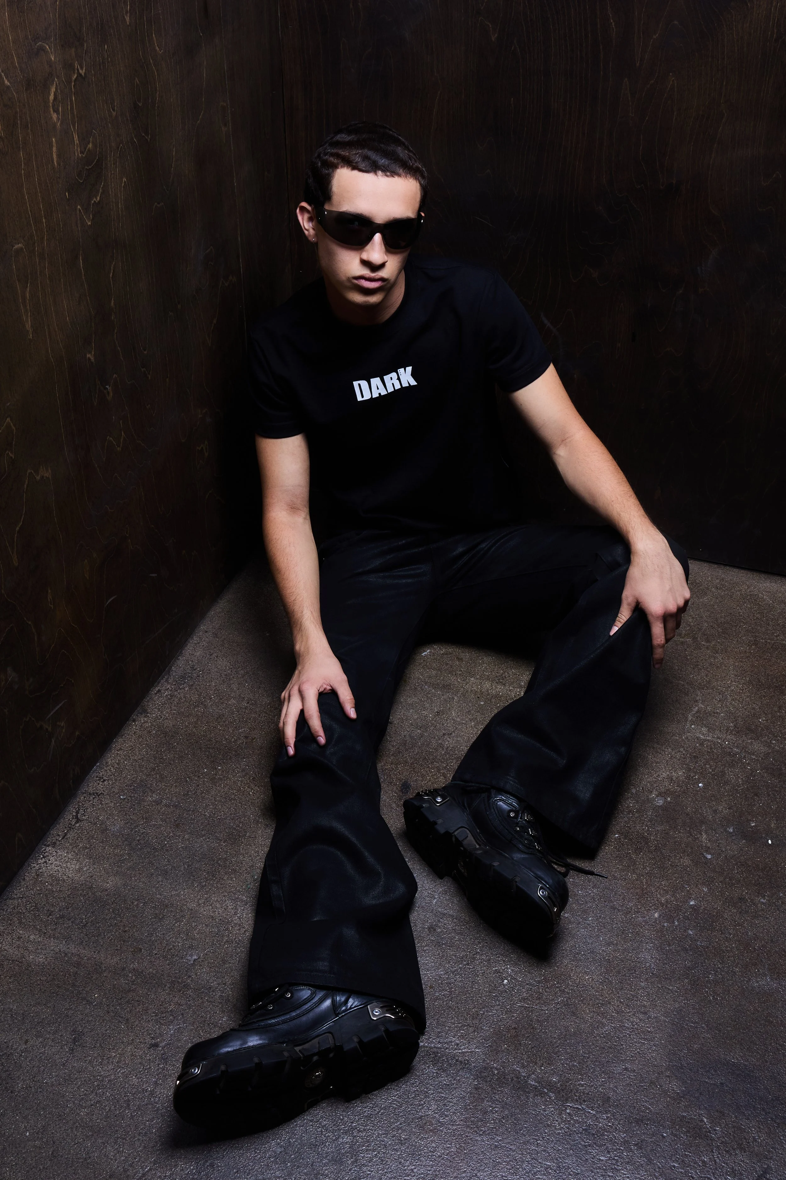 A young man wearing black clothing, sunglasses, and a T-shirt with the word 'DARK' written on it, sitting on the floor in a corner with dark wooden walls and a concrete floor.