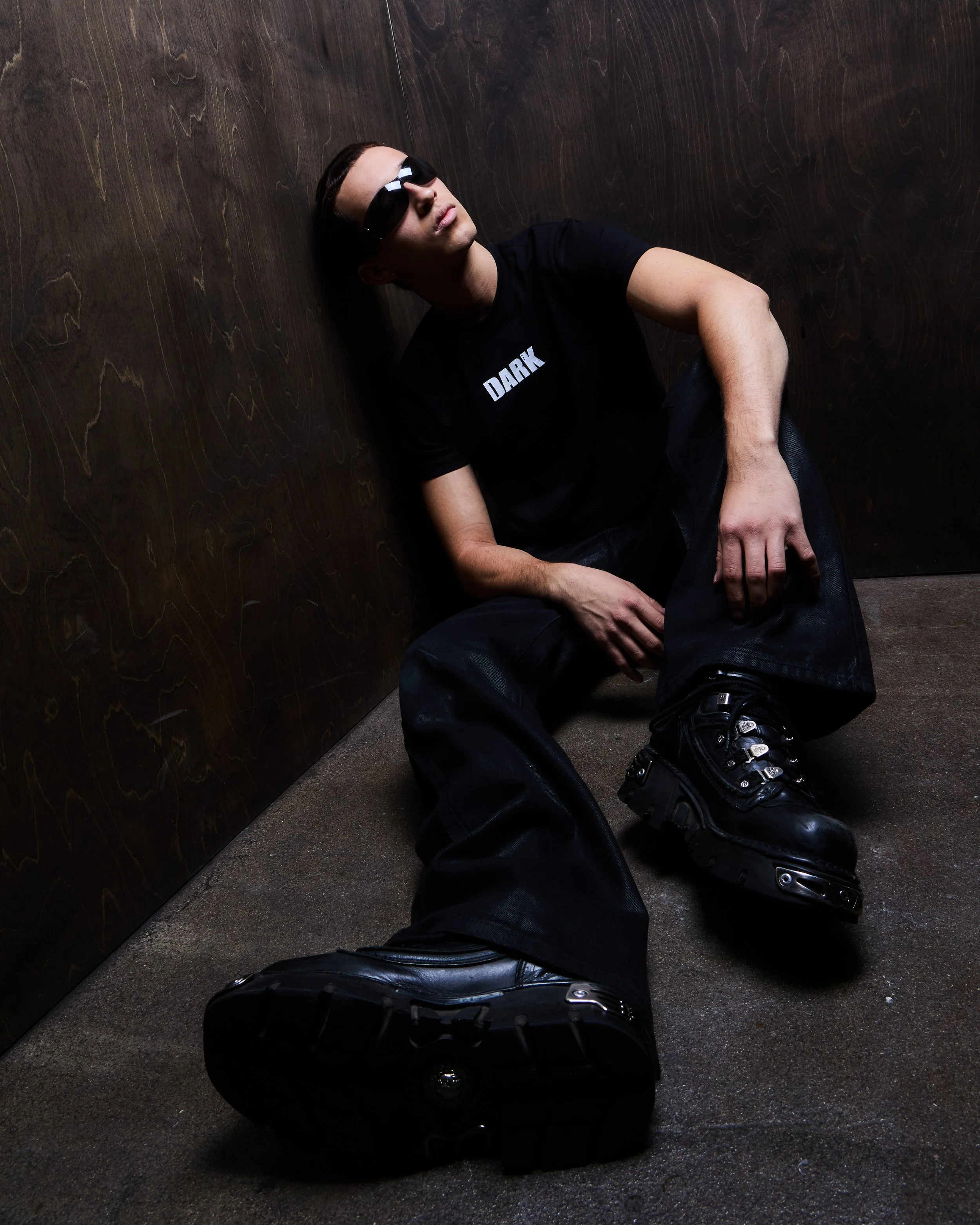 Young man with dark hair and sunglasses sitting against a dark wooden wall, wearing a black t-shirt with 'DARK' written on it, baggy black pants, and black boots, in a dimly lit room.