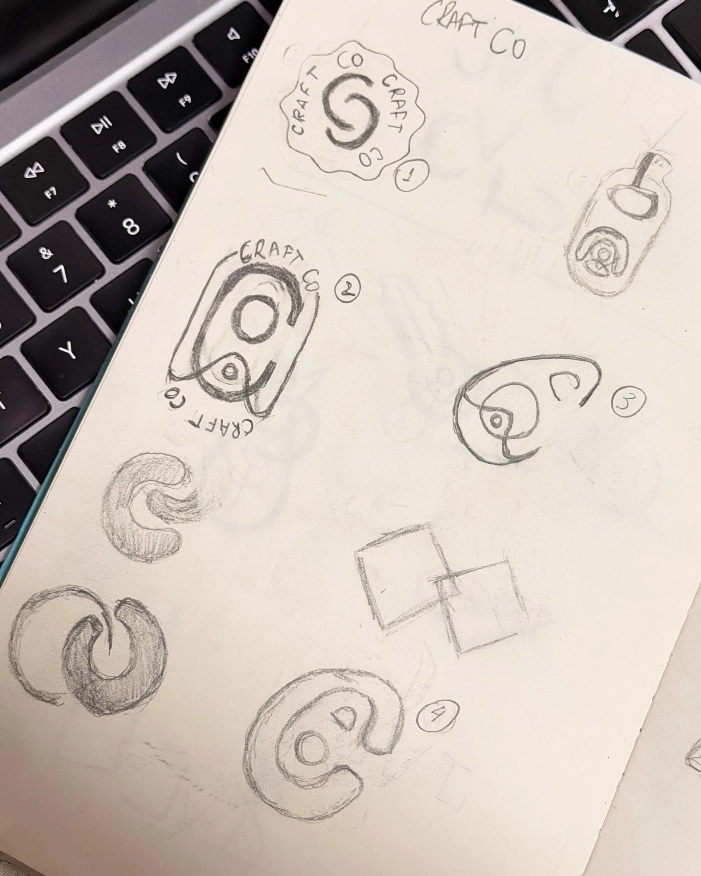 Sketches of various logo concepts for 'Craft Co' on a white sheet of paper, with some labeled and others shaded or outlined, positioned near a laptop keyboard.
