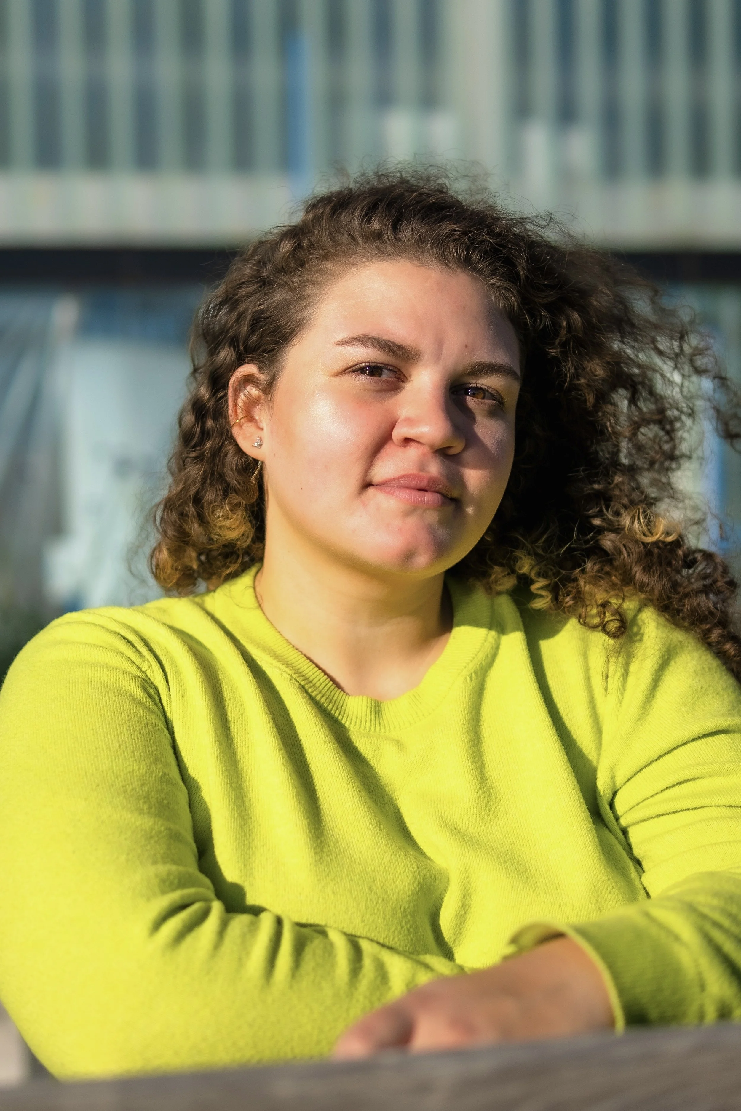 A young woman with curly brown hair and light skin, wearing a bright yellow sweater, sitting outdoors with a background of bookshelves or possibly library lockers, gazing attentively at the camera.