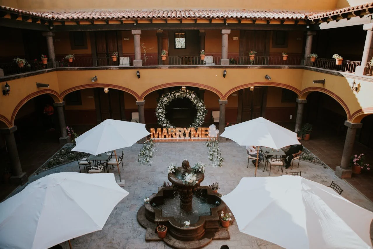 Indoor courtyard with tables and large white umbrellas, a fountain in the center, and a decorative floral arch with the letters "MARRYME" in front of the arch.
