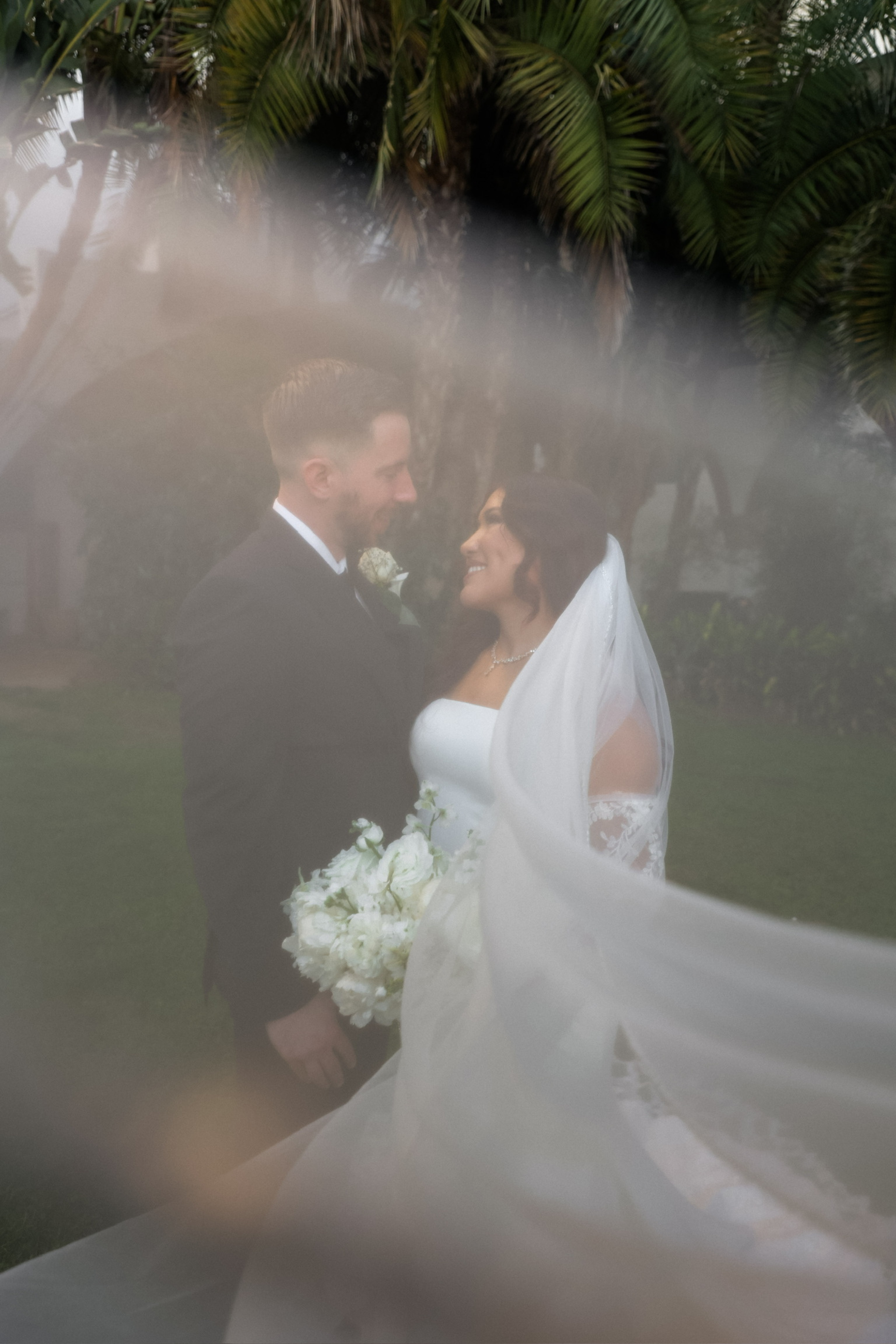 A bride and groom standing close together outdoors, smiling and facing each other, with greenery and palm trees in the background, through a semi-transparent veil.