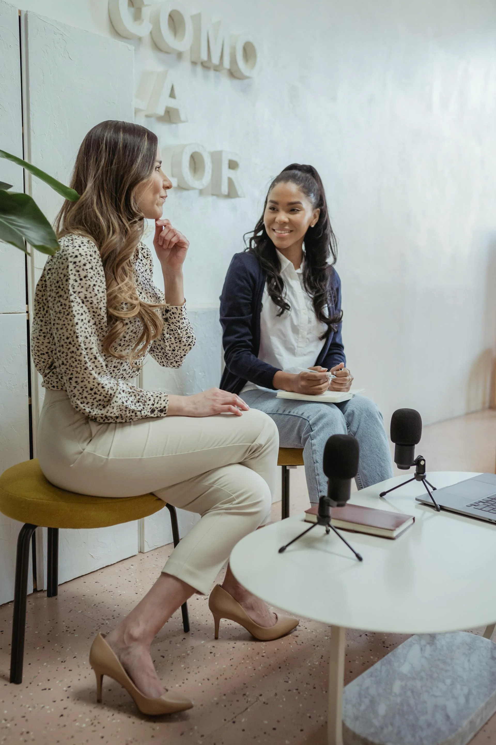 Two women engaged in a conversation during a podcast interview, seated in front of microphones, with a laptop and notebooks on a white table in a modern, well-lit room.
