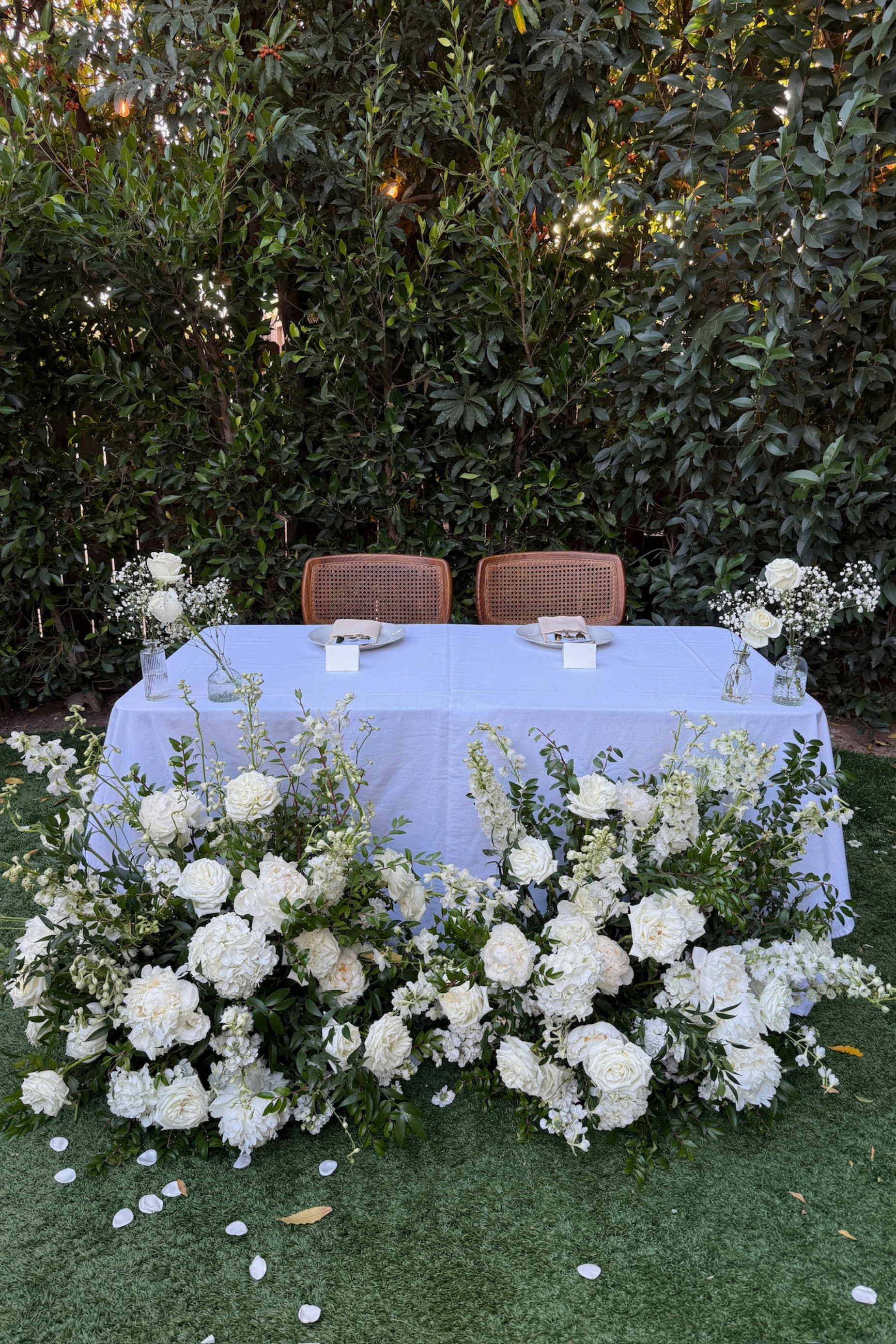 A decorated outdoor table with a white tablecloth, floral arrangements, and two chairs, set for a special occasion against a backdrop of green bushes.