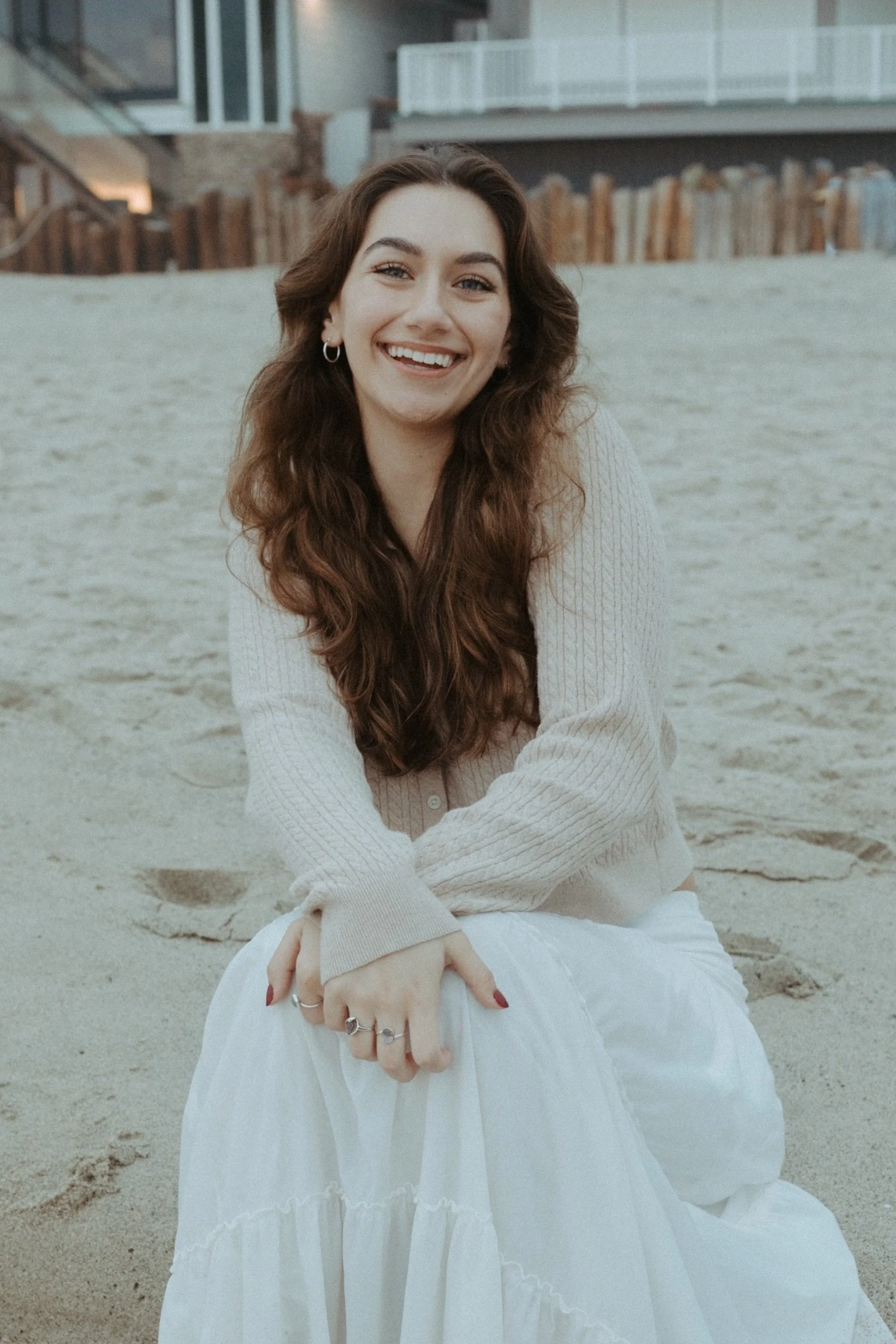 A young woman with long, wavy brown hair smiling while sitting on a sandy beach. She is wearing a cream-colored cardigan and a flowing white skirt.
