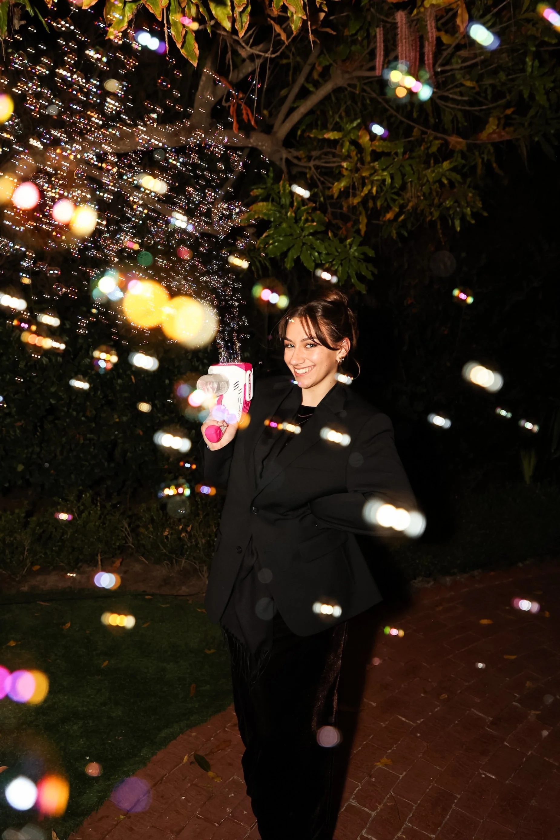 Woman in a black suit smiling and holding a bubble gun outdoors at night, with colorful bokeh lights and greenery around.