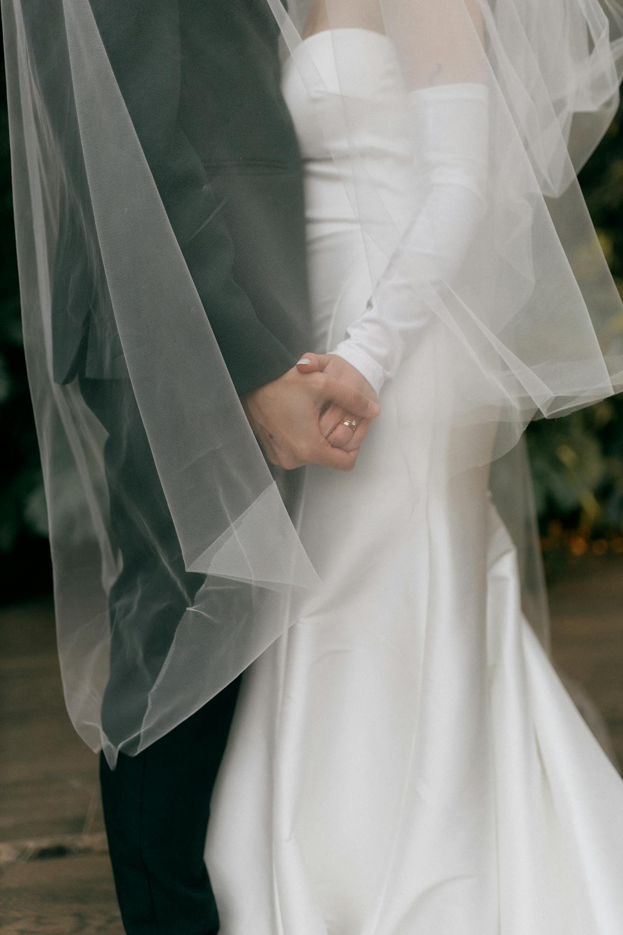 Close-up of a bride and groom holding hands, partly covered by a sheer veil, with the bride wearing a white dress and the groom in a dark suit.