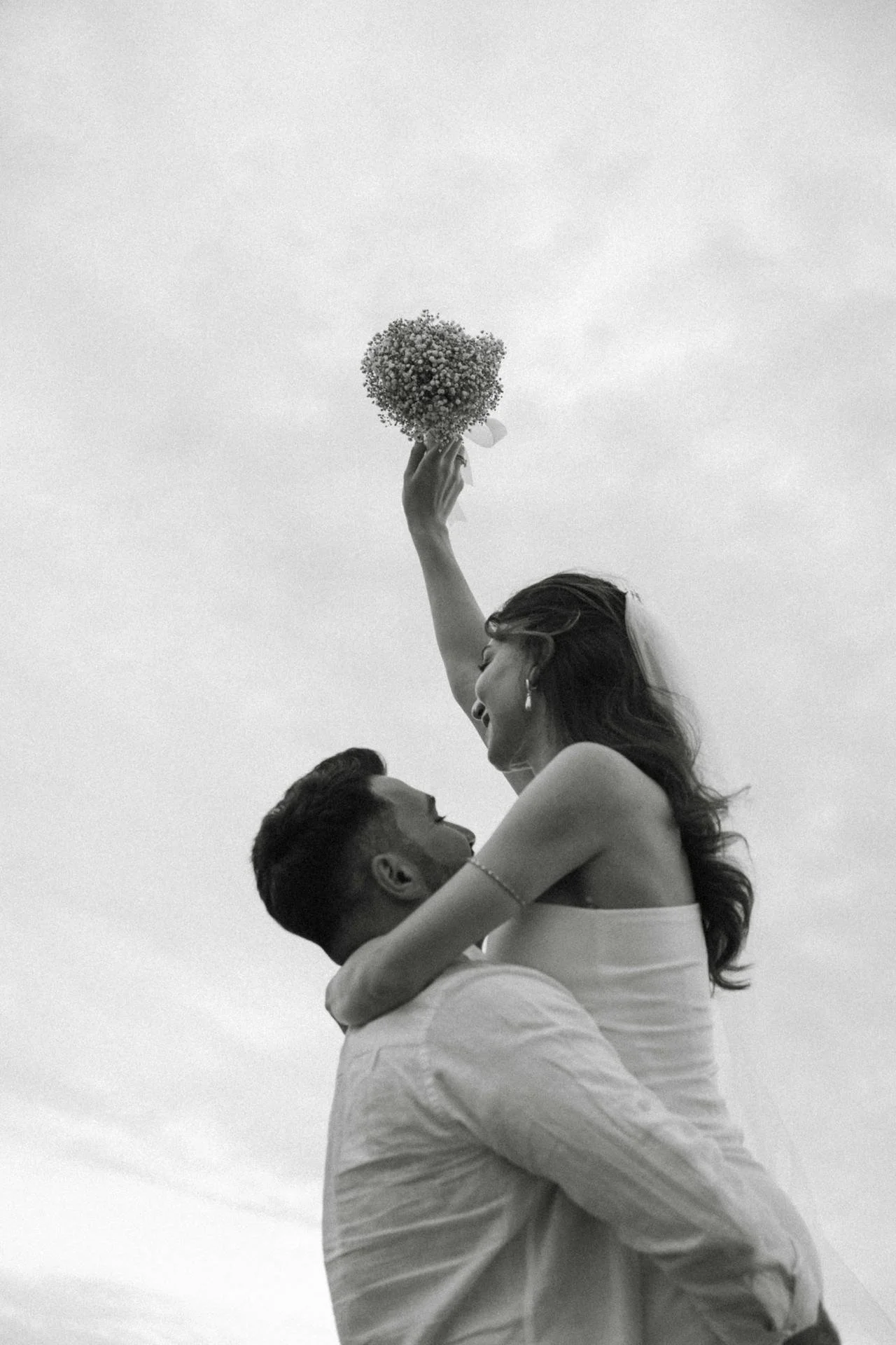 A black-and-white photo of a couple, with the woman sitting on the man's shoulders, raising her arm with a bouquet of flowers, smiling.