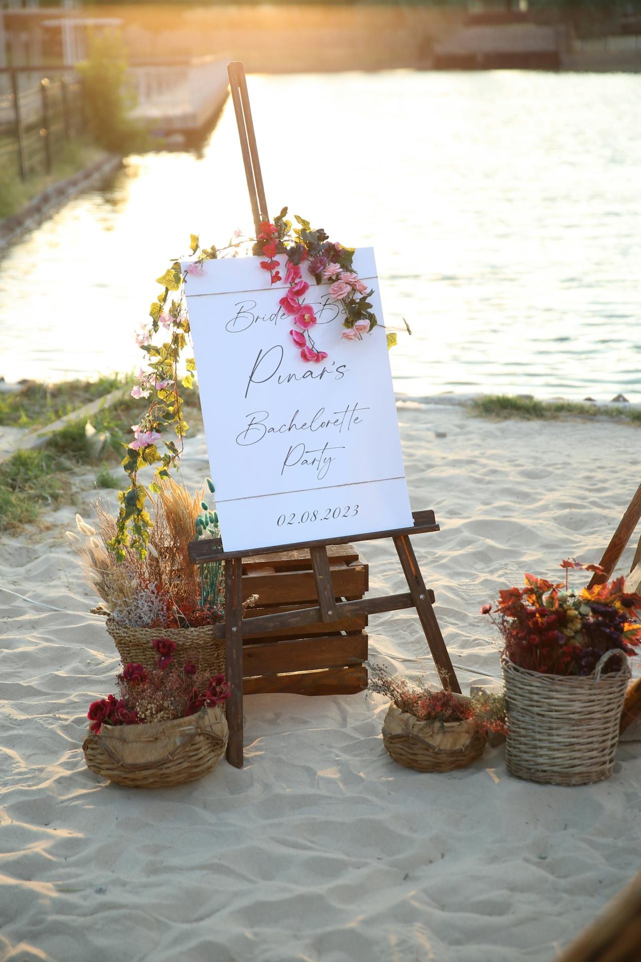 A wedding sign on an easel on the beach reads 'Bride Pinard's Bachelorette Party' with the date 02.08.2023, decorated with pink and purple flowers, surrounded by baskets of flowers on sandy ground, with water and a sunset in the background.