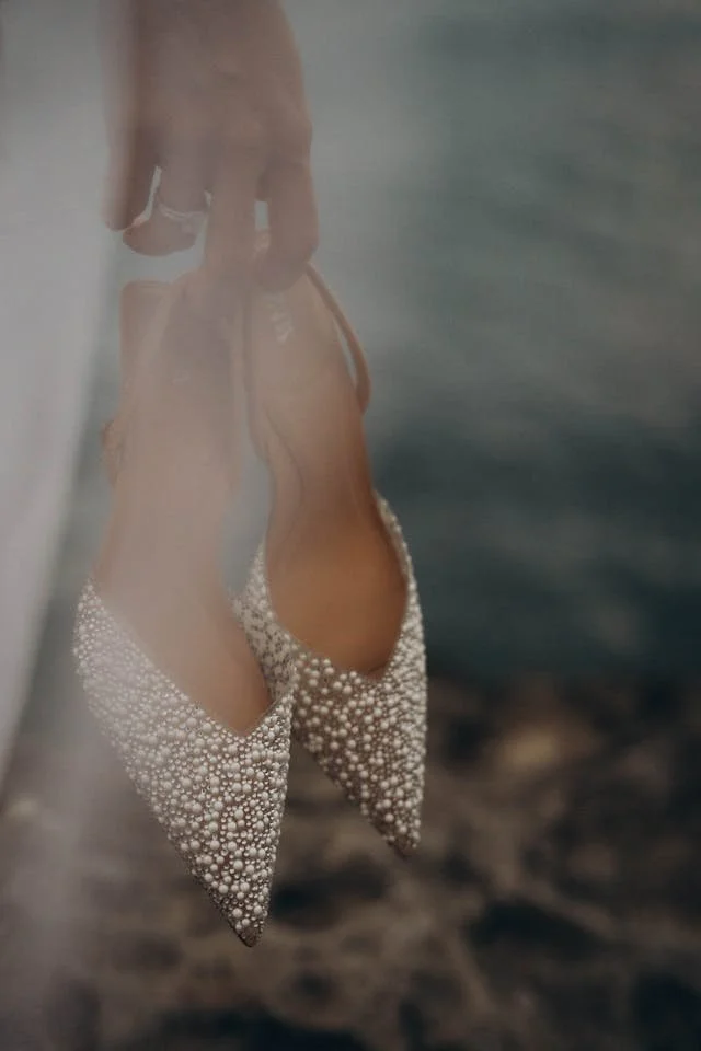 Pair of beige high-heeled shoes with pearl embellishments hanging on a person's finger.