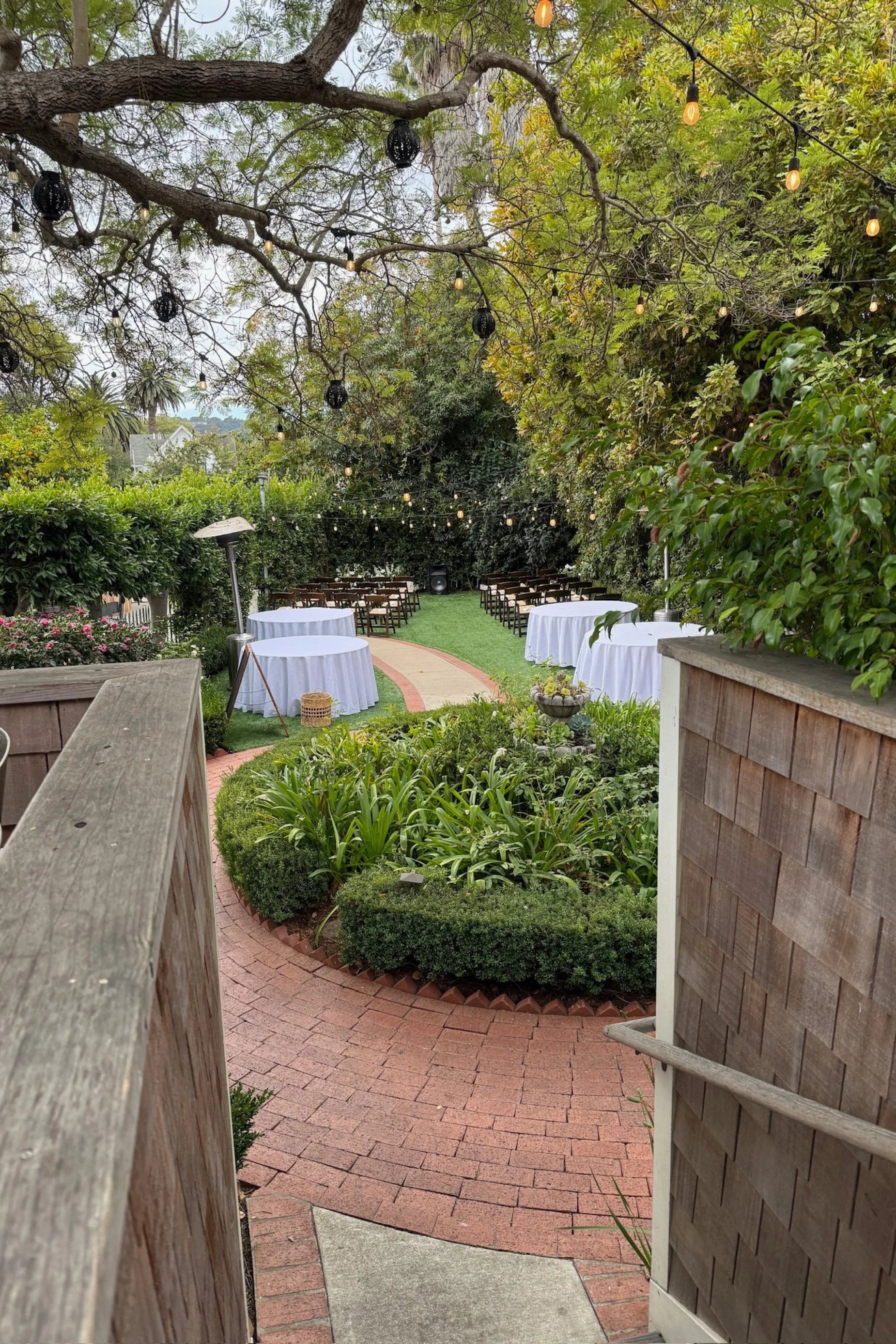 An outdoor garden setting prepared for a gathering or event with white-draped tables, chairs, string lights hanging from trees, and a path leading to the seating area.