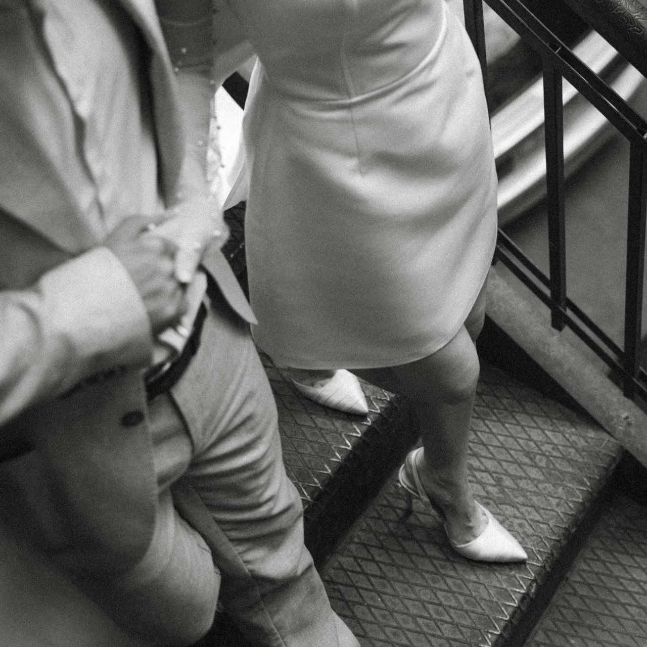 Black and white photo of two people standing on a metal staircase, captured from above. One person is wearing light-colored pants with a buttoned shirt, and the other is wearing a light-colored dress and pointed shoes.
