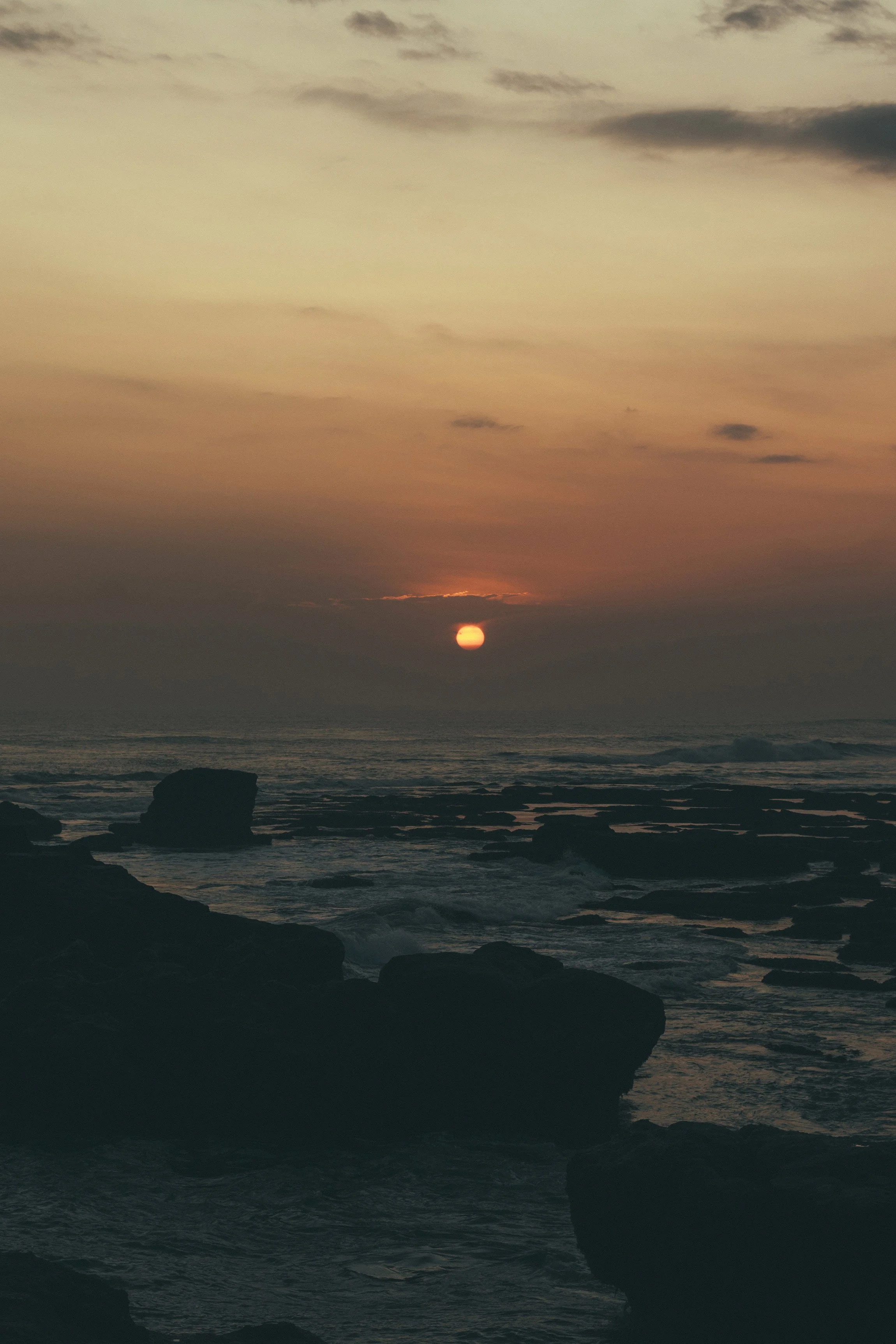 Sunset over the ocean with rocks in the foreground and a cloudy sky.