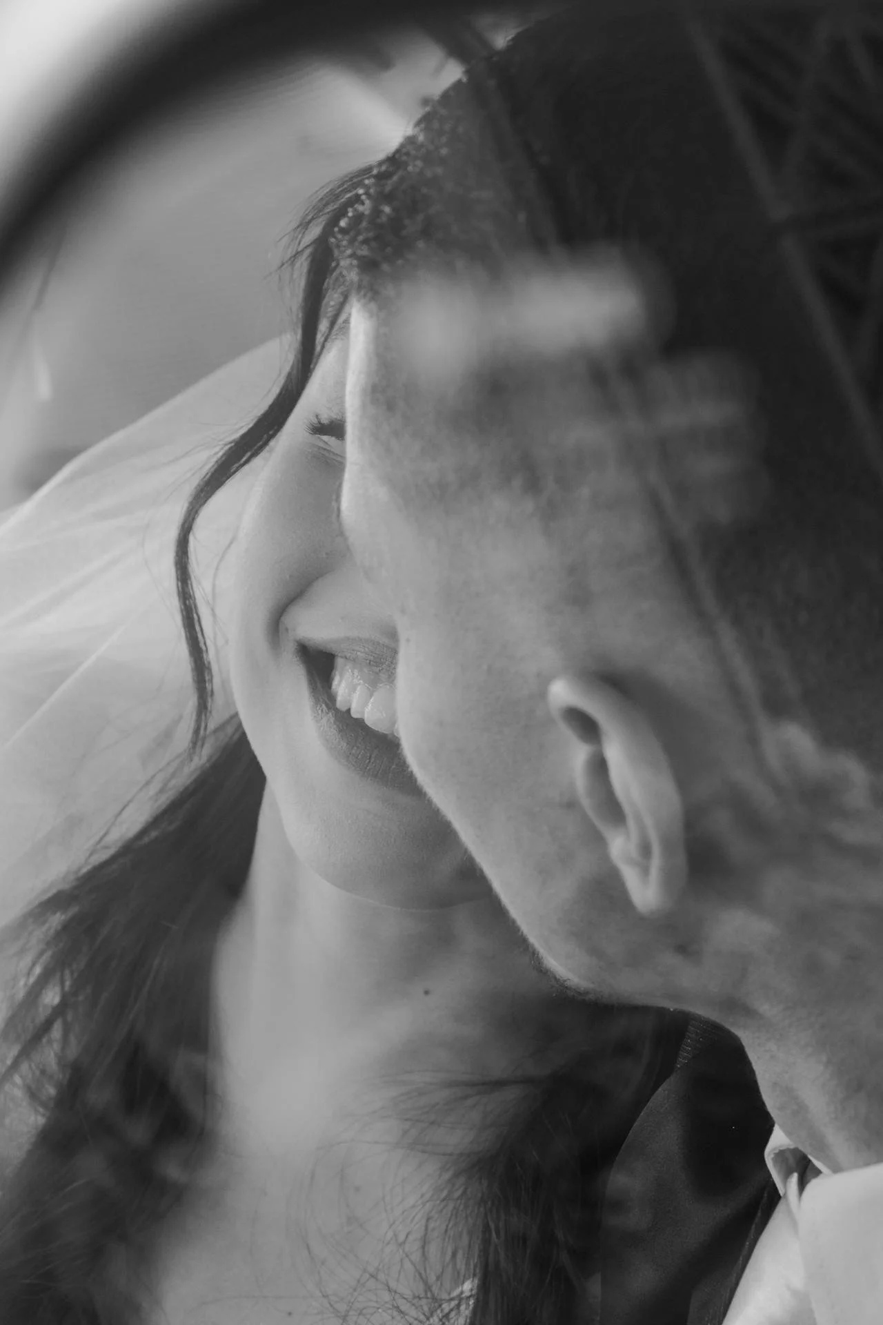A close-up black and white photo of a smiling woman and a man face to face, almost touching noses, with their eyes closed, in an intimate moment.