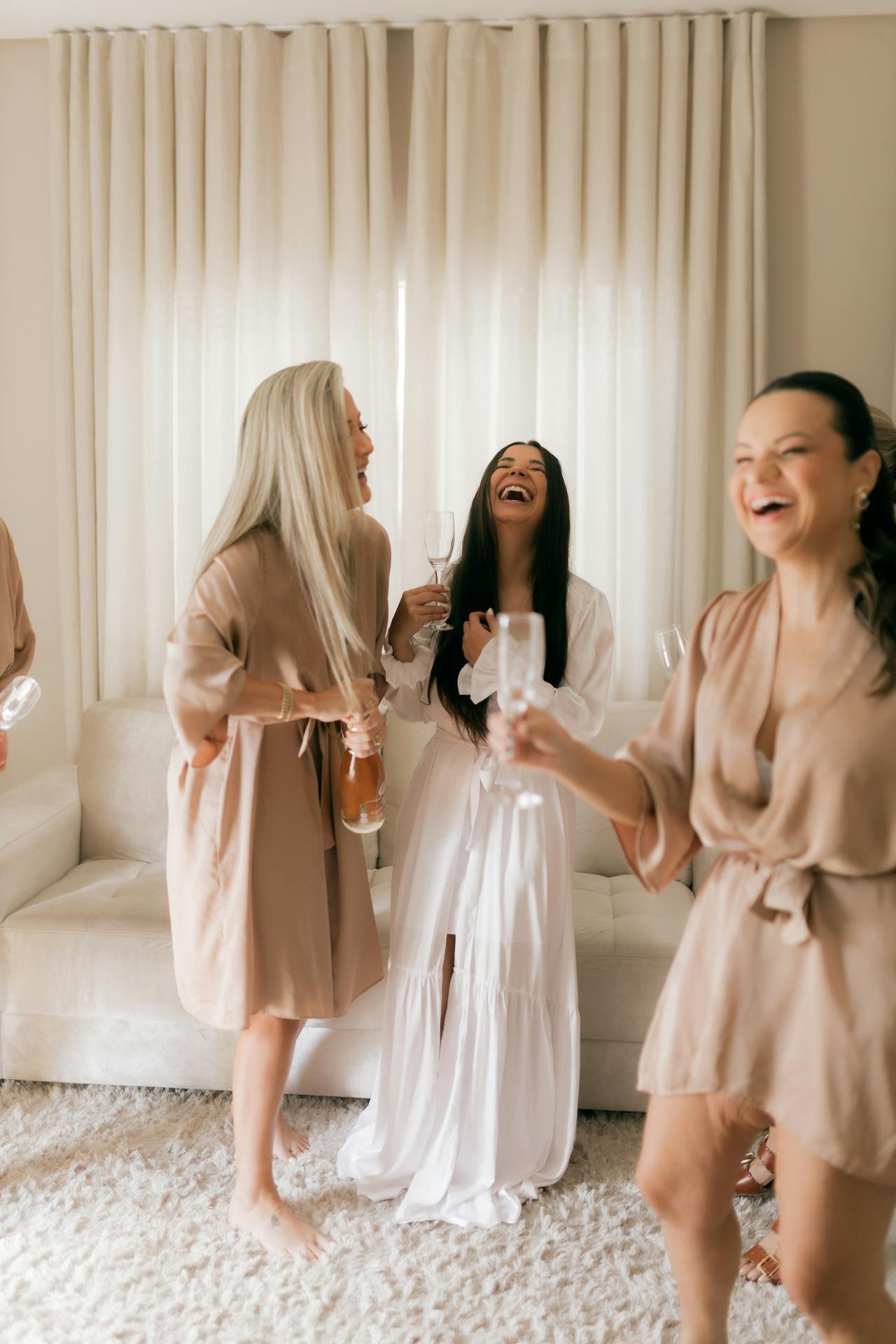 Group of women in beige and white dresses laughing and celebrating with champagne in a well-lit living room.